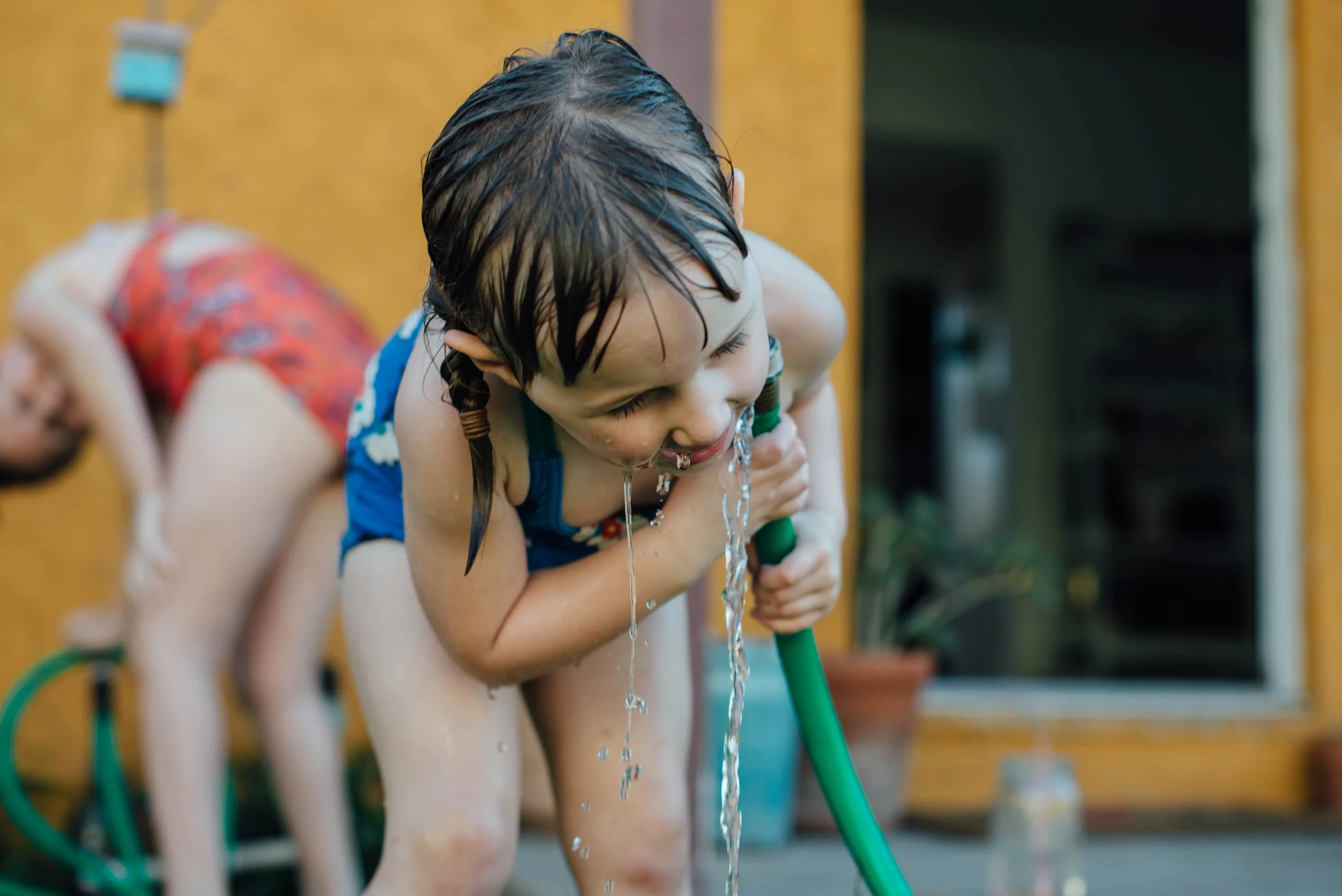 A young boy with wet hair wearing a swimsuit, holding a green garden hose, leaning forward and laughing as water sprays out.