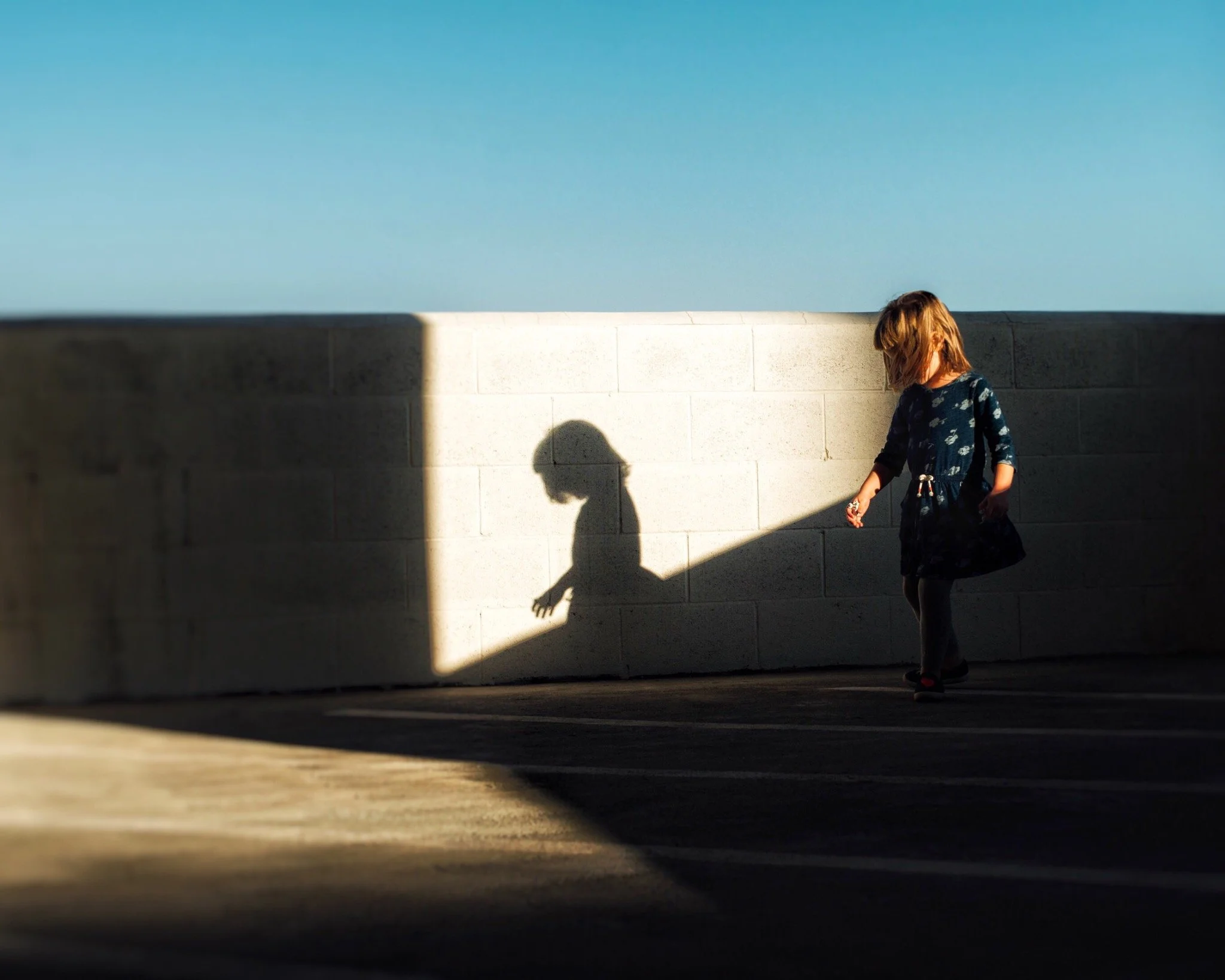 A young girl with brown hair, wearing a blue dress with white patterns, is walking next to a light-colored cinder block wall under a clear blue sky. Her shadow is cast on the wall.