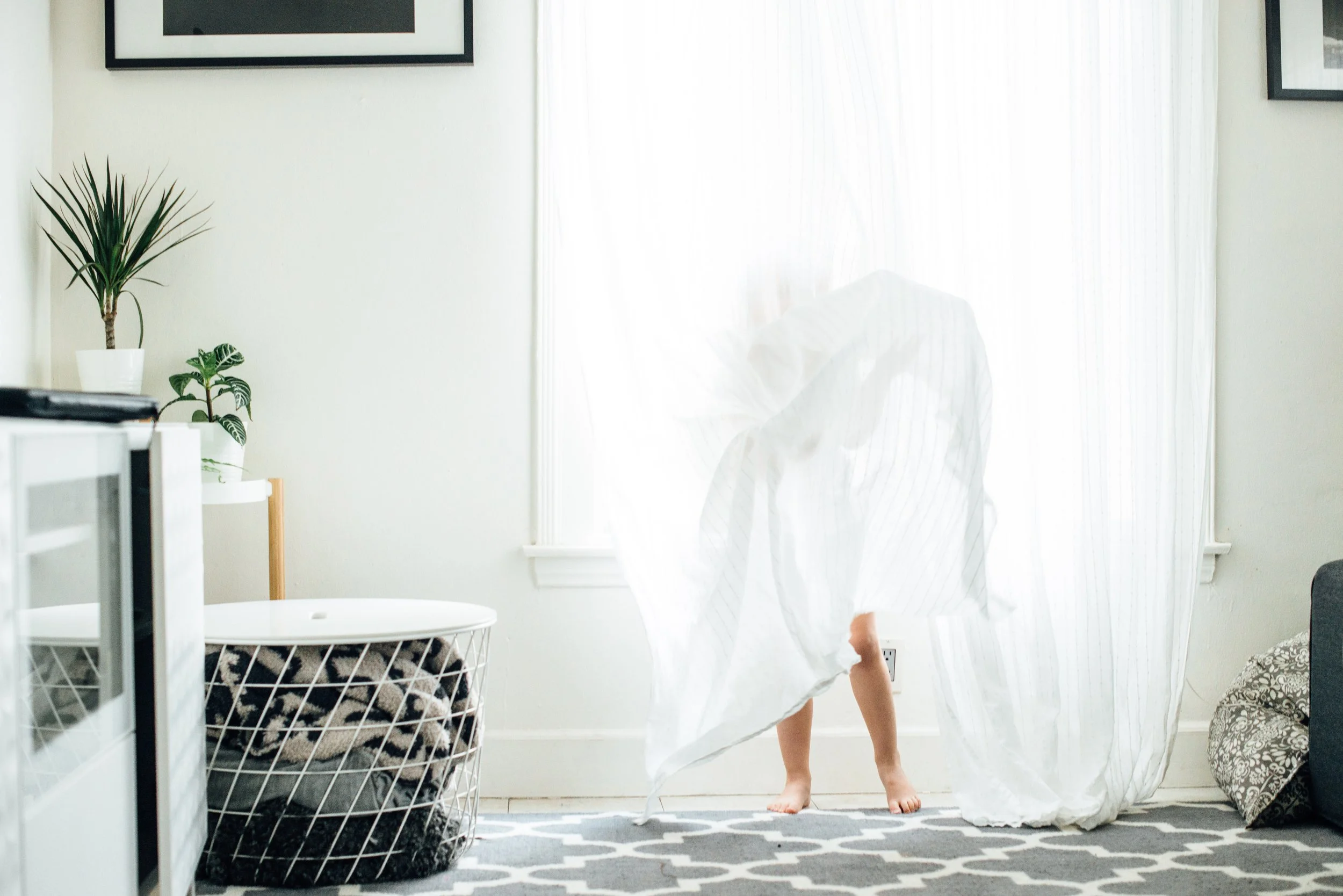 A person in a white dress is walking across a room illuminated by sunlight through sheer curtains. The room has a black-and-white patterned rug, white walls, potted plants, and framed pictures hanging on the wall.