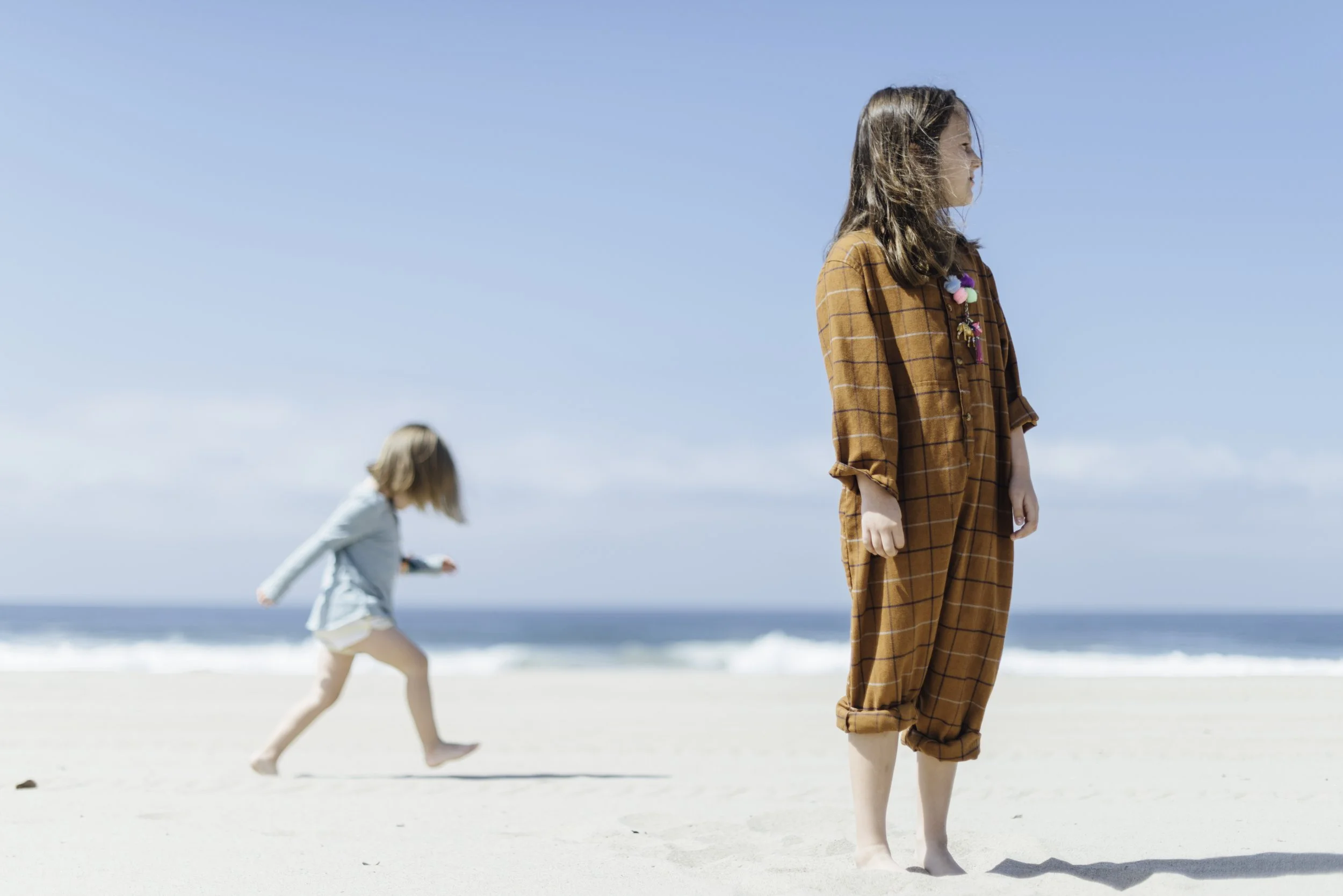 A young girl in a brown checkered outfit standing on a sandy beach, with other children playing near the water in the background under a clear blue sky.