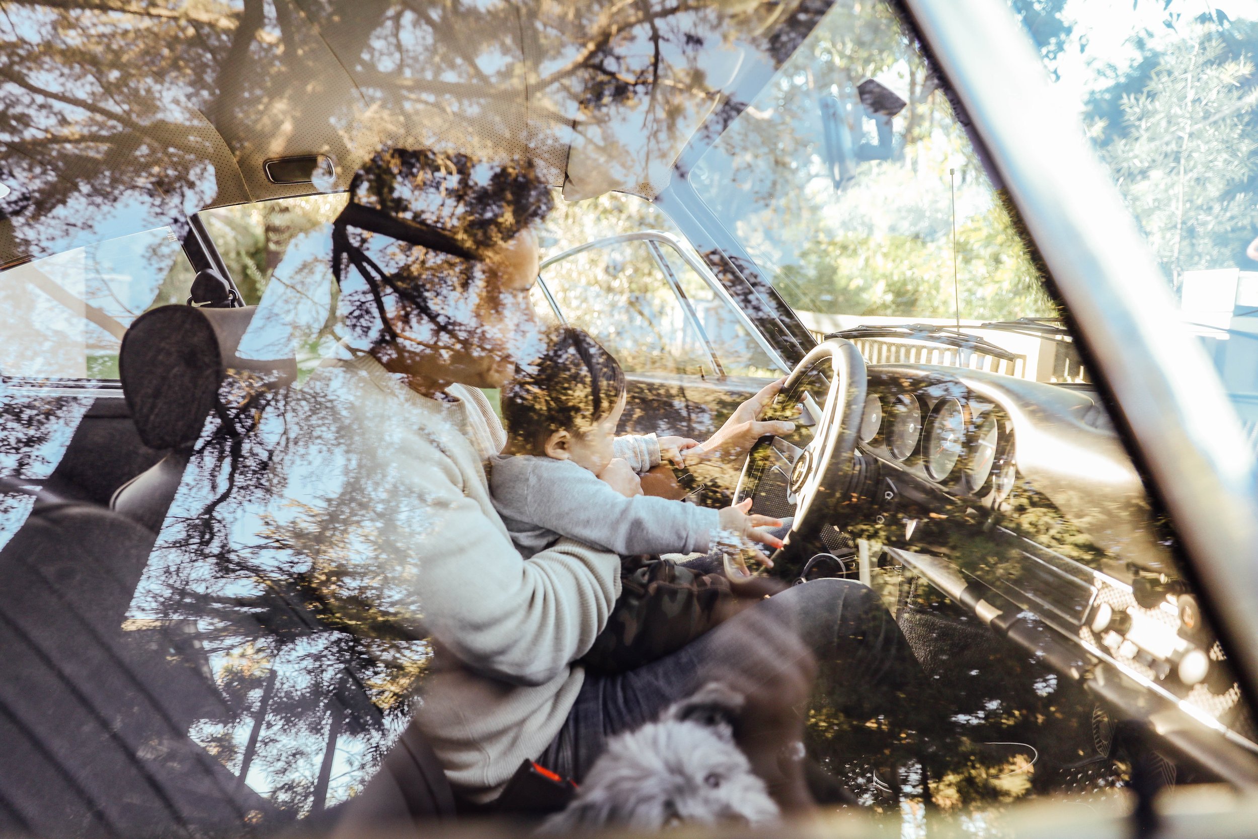 View from inside a car showing a woman and a child in the front seats, with reflections of trees on the windshield, sunlight, and the dashboard visible.