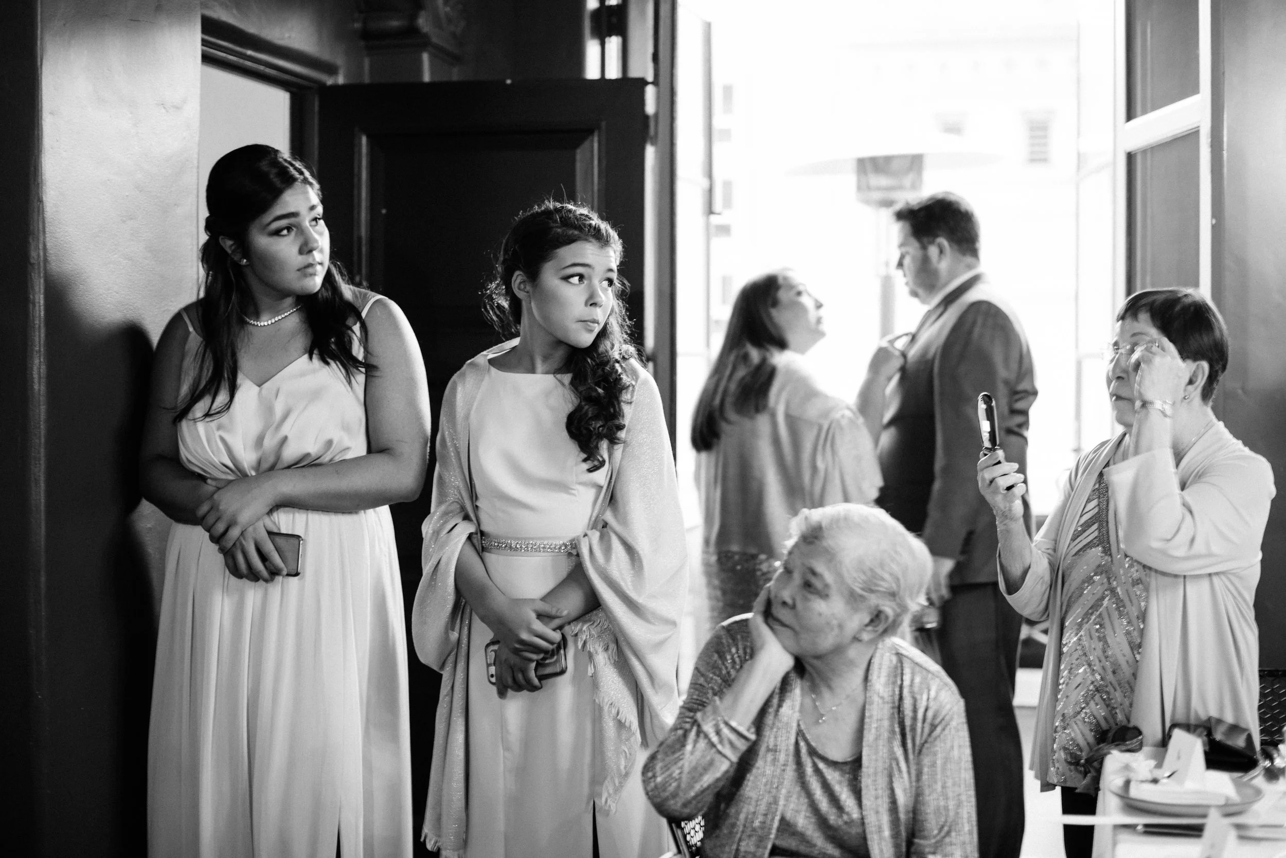 Two young women in formal dresses standing by a wall, looking in opposite directions, with one holding a phone. In the background, an older woman looking at her phone, a young man in a suit, and an elderly woman sitting at a table are visible. The sc