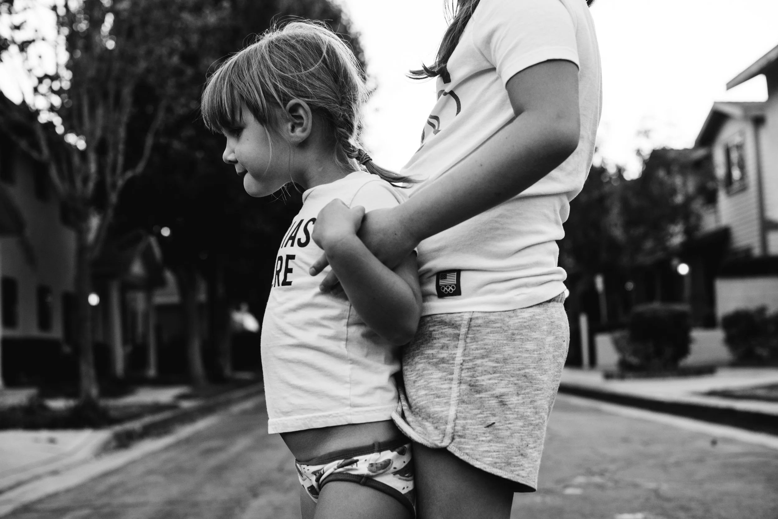 A young girl and an older girl or woman, possibly an adult, embrace outdoors. The young girl is wearing a t-shirt and underwear, and the older girl or woman is wearing shorts and a t-shirt. Both are standing close, holding hands with the young girl l