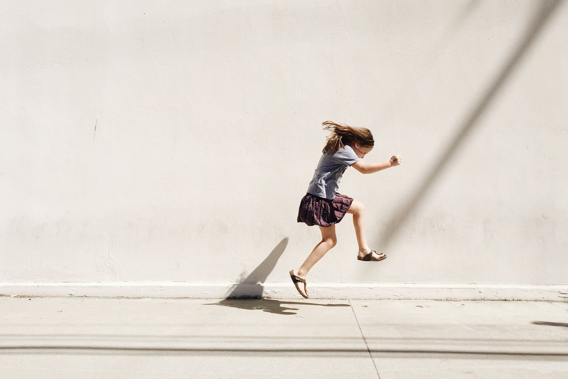 A young girl with long hair running or jumping in front of a plain beige wall, casting a shadow on the ground.