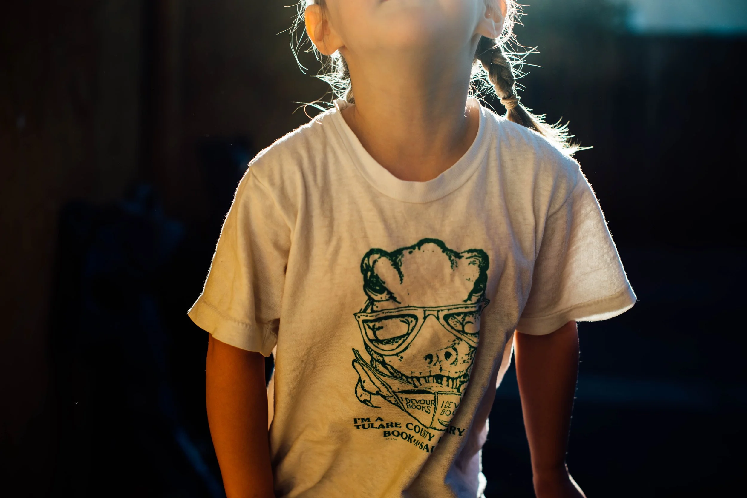 Close-up of a child's torso wearing a white T-shirt with a graphic of a bear wearing sunglasses and text, with sunlight highlighting the child's hair and ear.