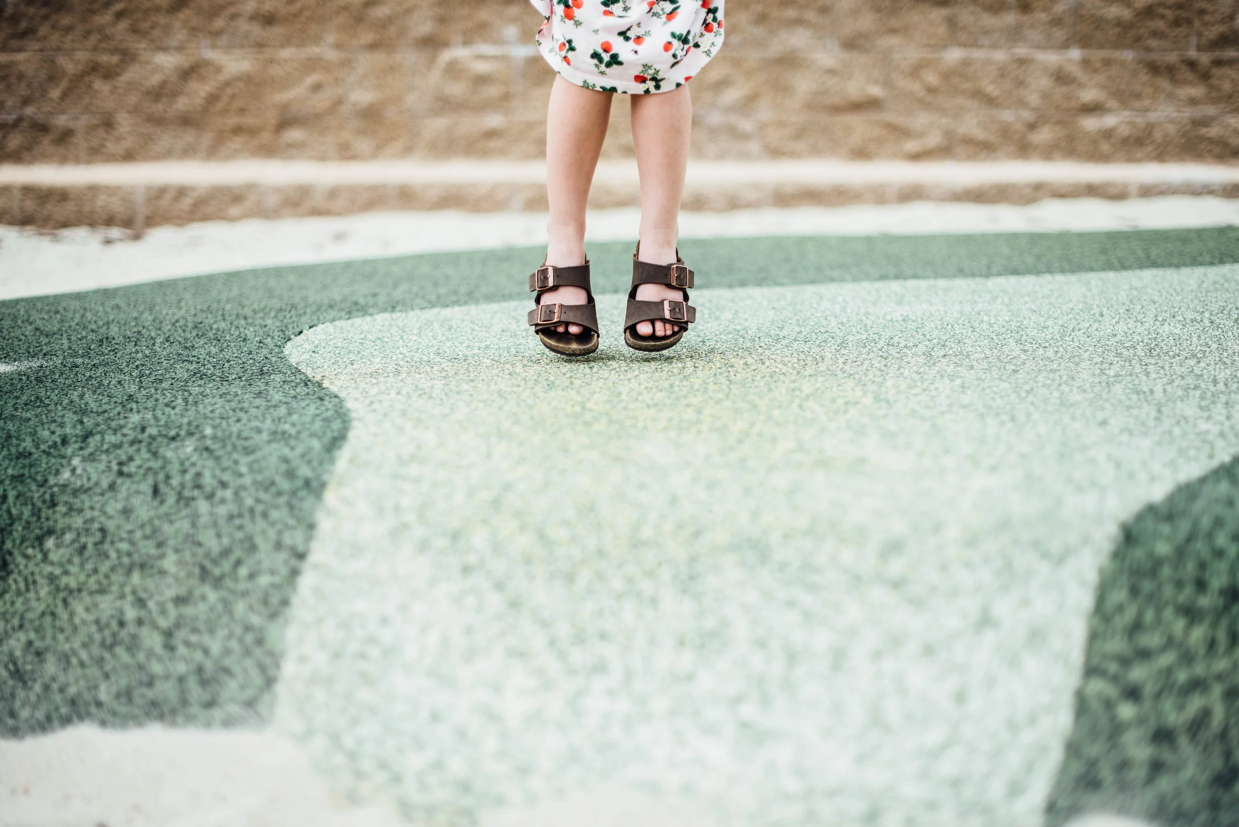 A child's legs and feet wearing brown sandals standing on a colorful painted surface outdoors.