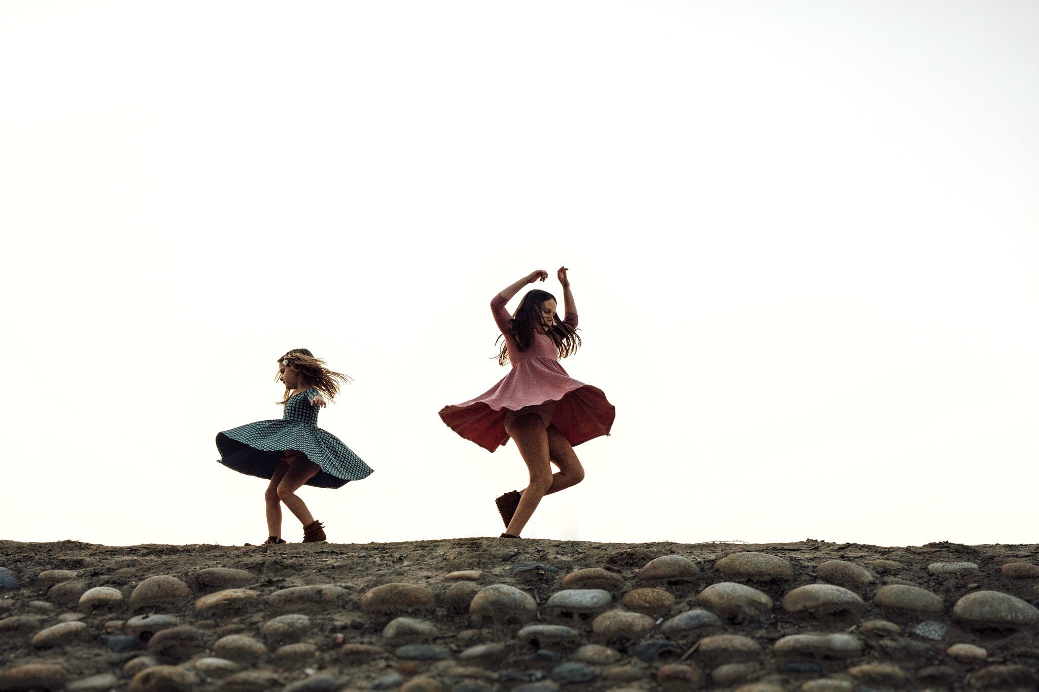 Two girls in dresses twirling on rocky ground with a plain sky background.