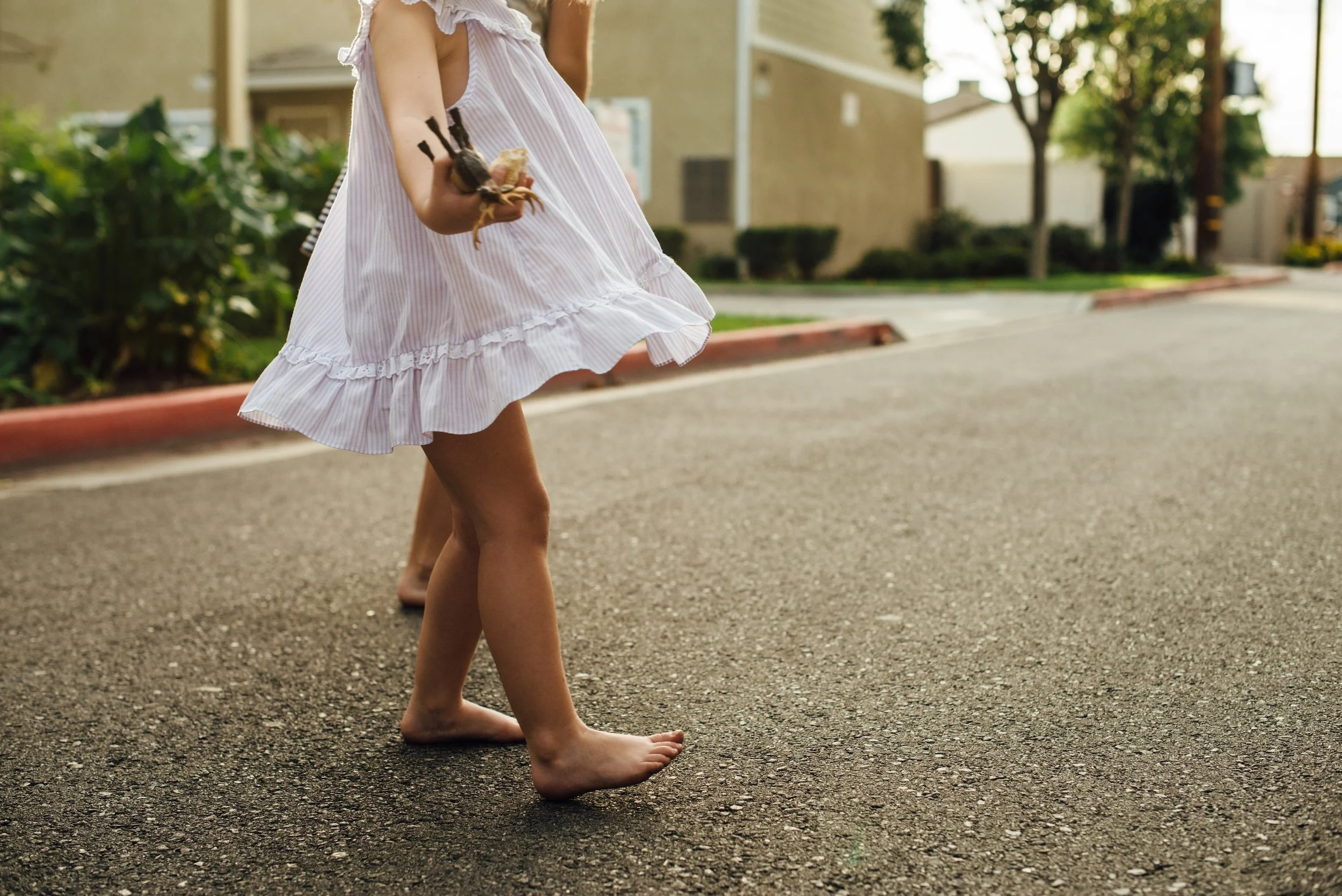 A young girl walking barefoot on the street carrying a small spider in her hand, wearing a light, pink and white striped summer dress with ruffles.