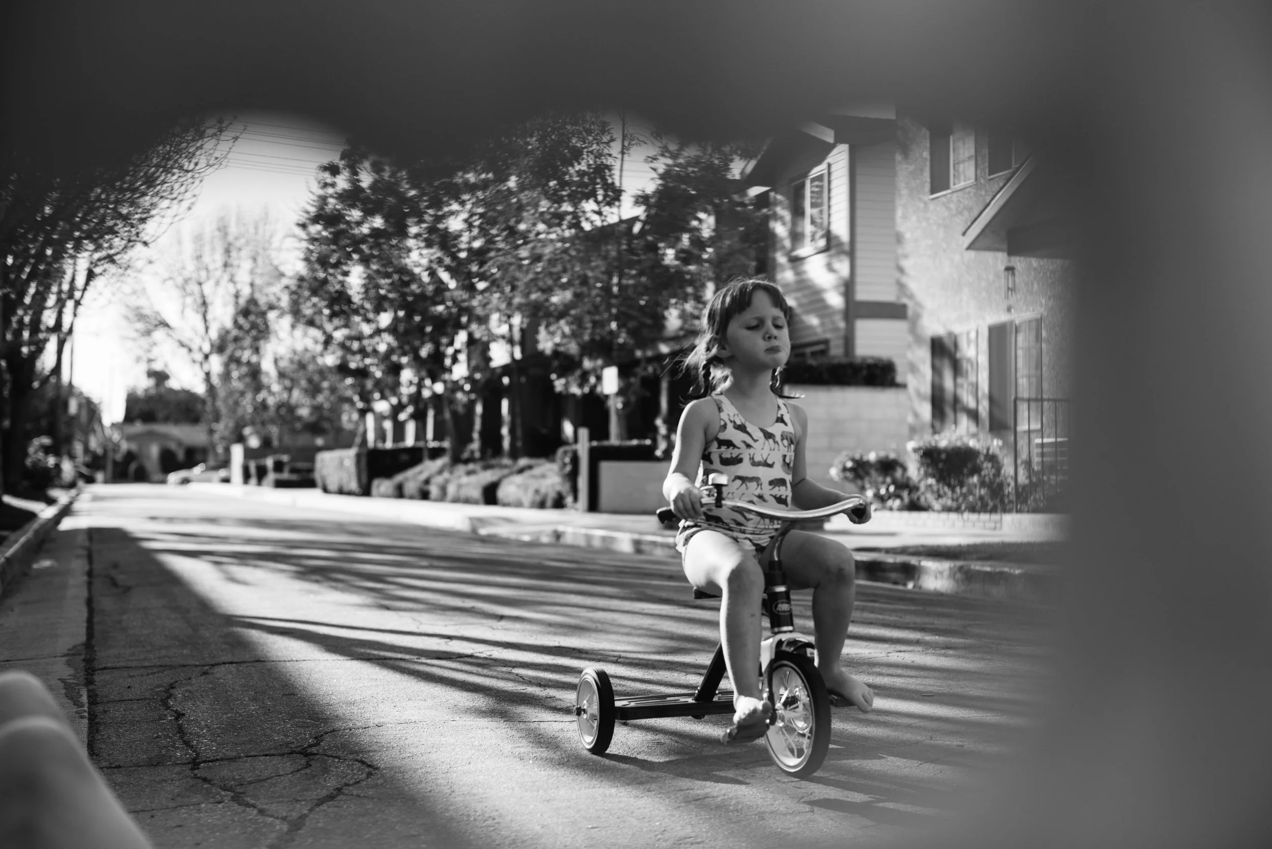 A black-and-white photo of a young girl riding a tricycle on a residential street. She is wearing a sleeveless top with a cat pattern and has her hair tied back. The street has trees, houses, and a sidewalk, with her being partially framed by an out-
