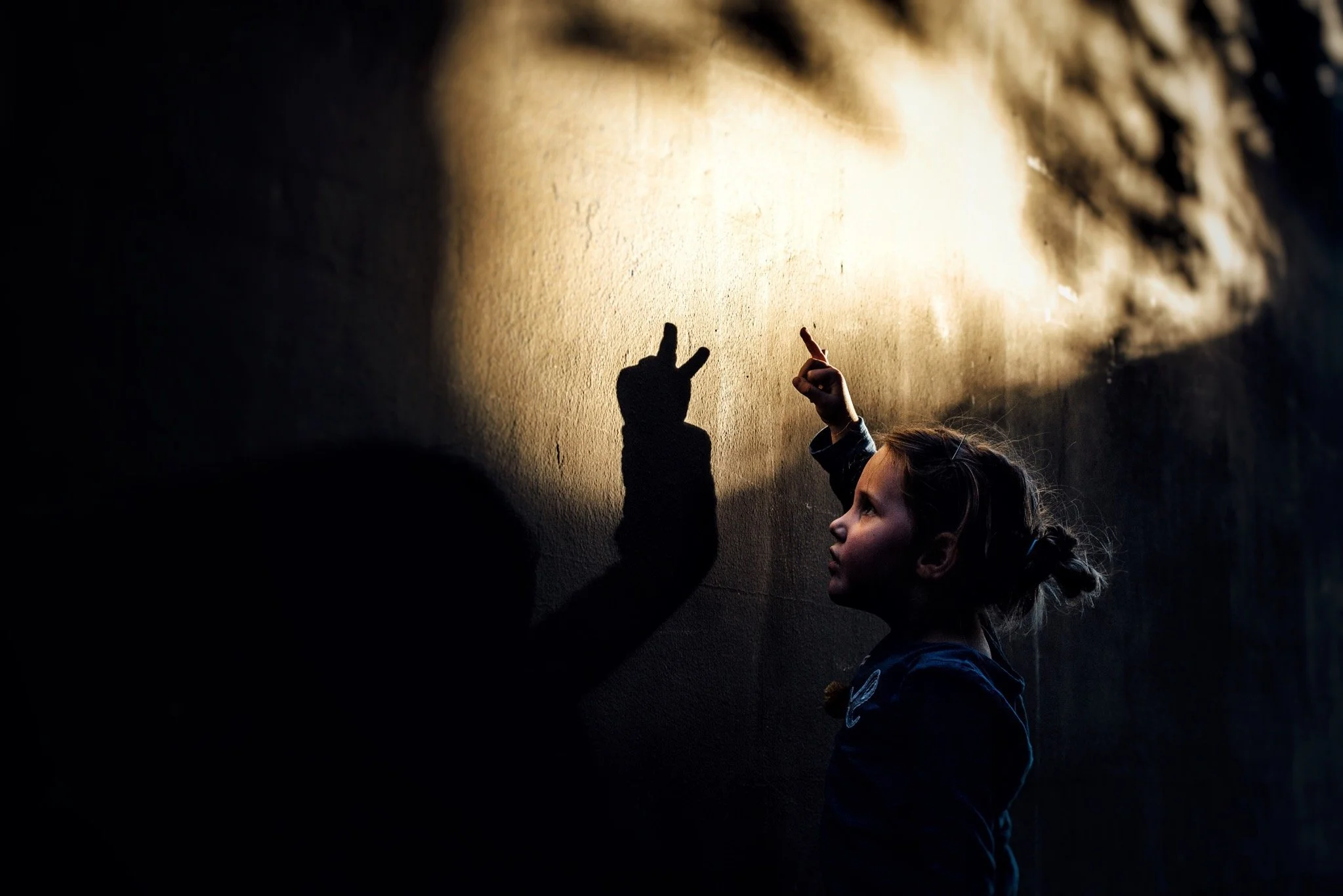 A young girl with blonde hair tied in a bun faces a sunlit wall, flashing a peace sign with her right hand.