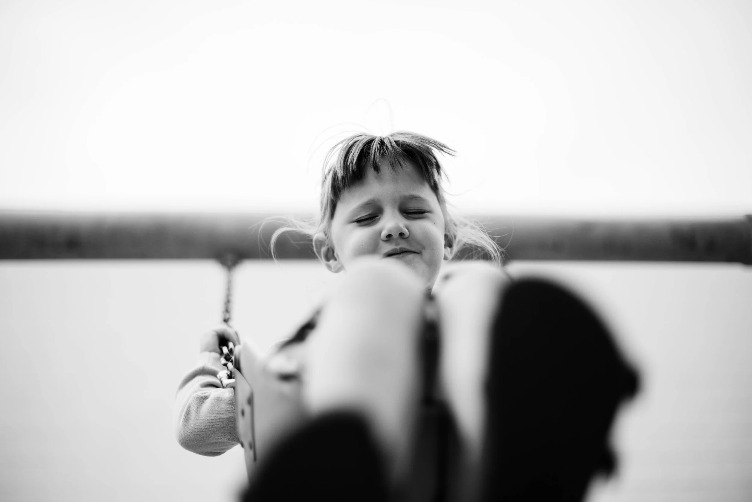 A young girl with short hair and a closed-eye expression, looking downward while holding onto a swing or similar object, with a blurred landscape and sky in the background.