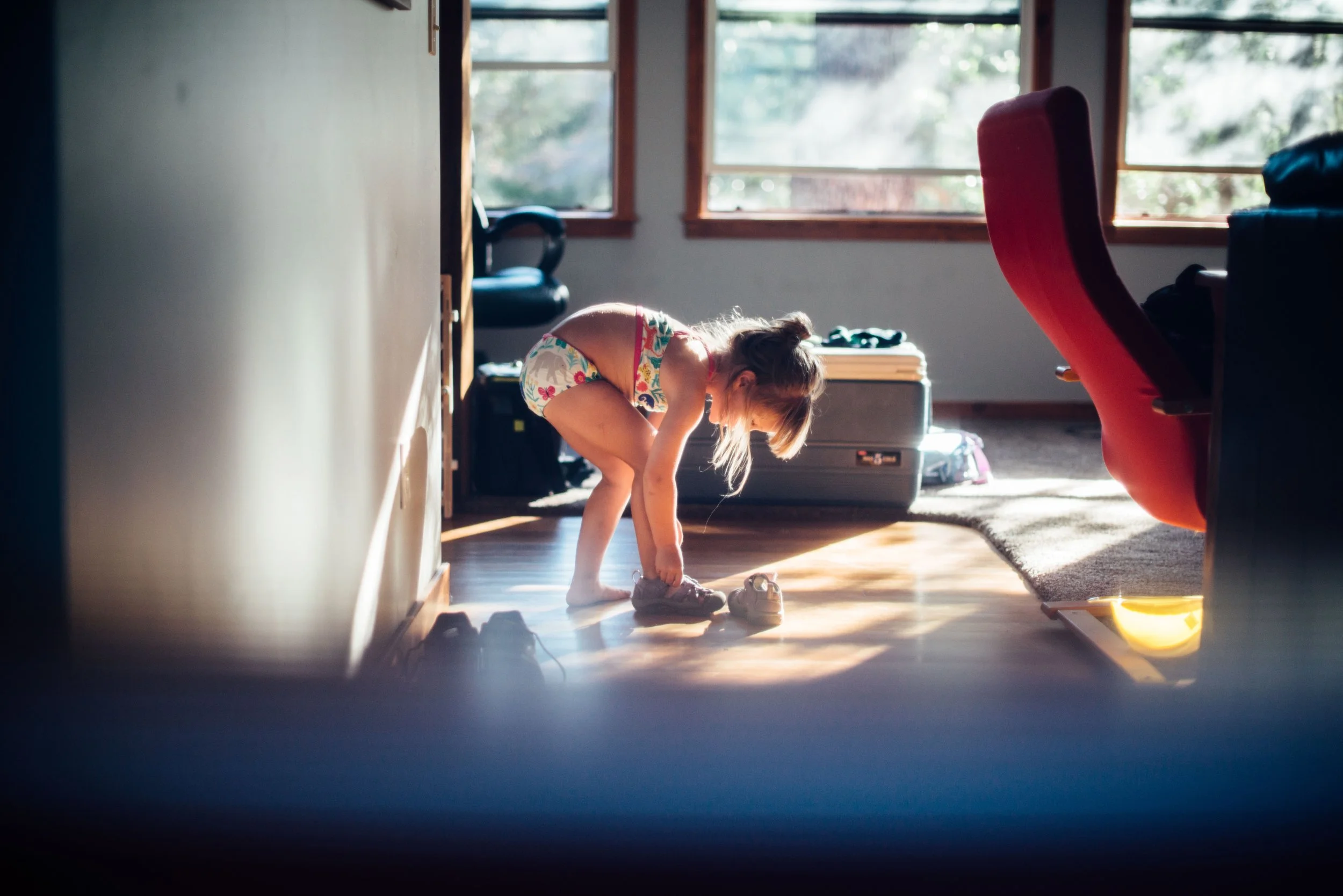 A young girl crouches down in a sunlit room, holding her shoes. She is wearing colorful, patterned shorts and a top with floral all-over print. The room has large windows with trees visible outside, and various objects, including a red chair and a su
