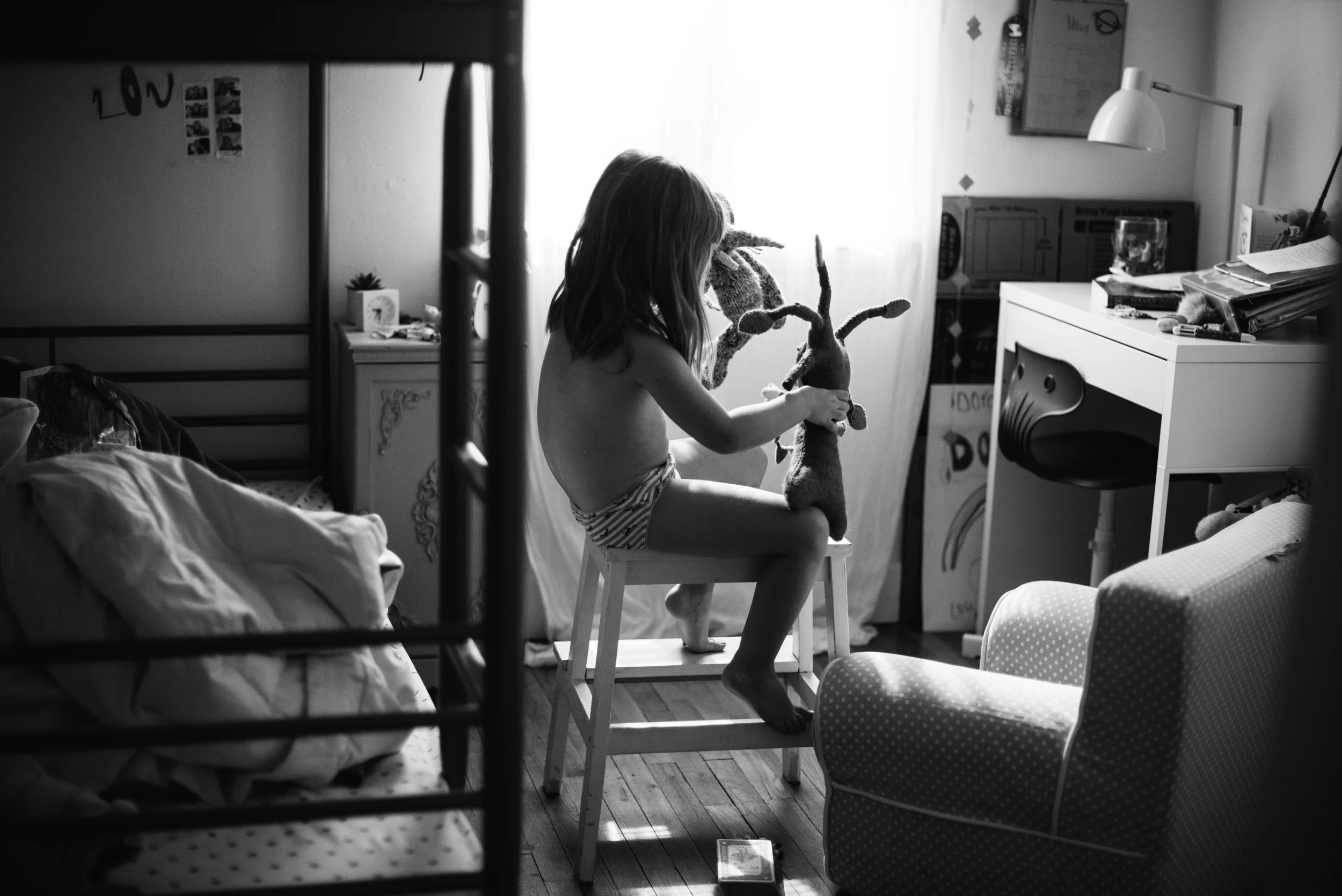 A young girl with shoulder-length hair, sitting on a small stool, holding a stuffed reindeer toy inside a room with a cluttered desk, a white dresser, and a bed. The room is illuminated by natural light from a window.