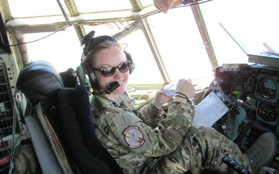 A smiling woman in military uniform sitting in the cockpit of an aircraft, wearing sunglasses and a headset.