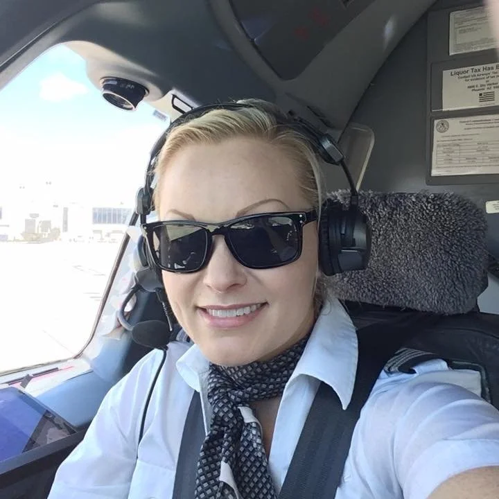 A professional female pilot wearing sunglasses, a headset, and a uniform, sitting in the cockpit of an airplane with a bright sky outside.