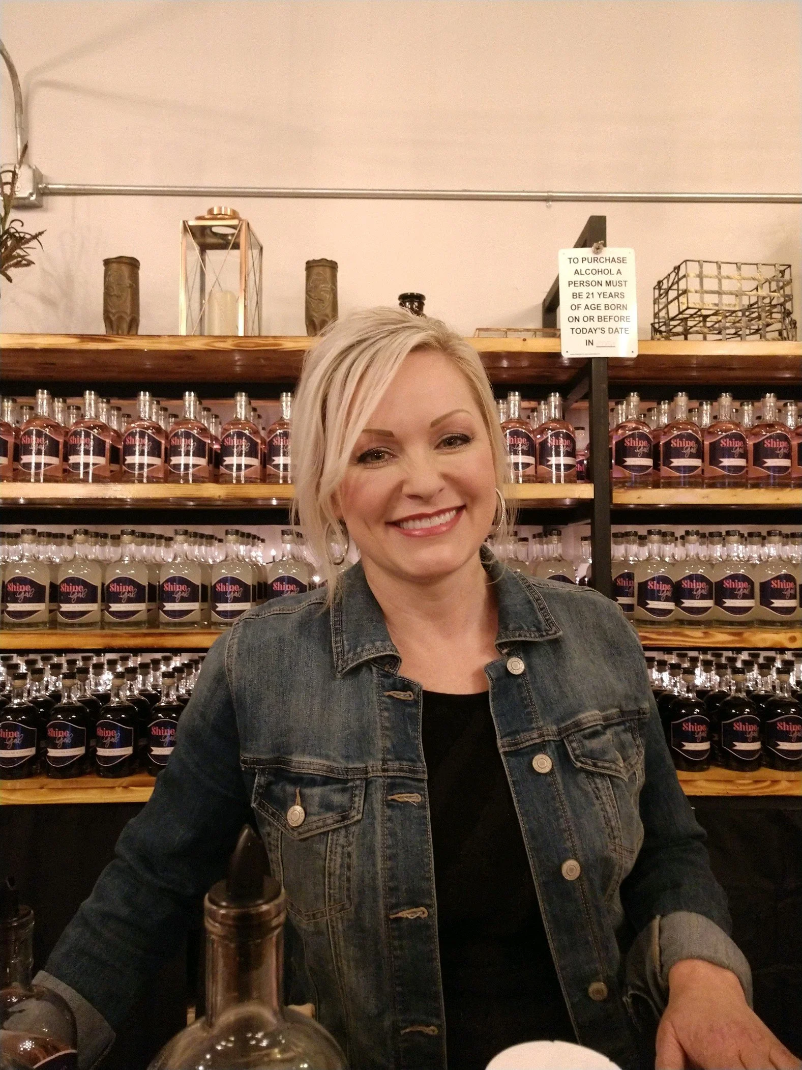 A woman with blonde hair smiling in front of shelves lined with bottles labeled Shine.