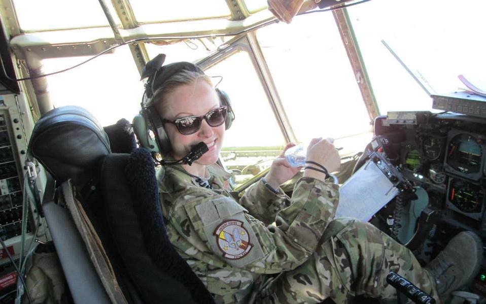 A woman in military uniform sitting in the cockpit of an aircraft, wearing sunglasses and headsets, holding a water bottle, smiling.