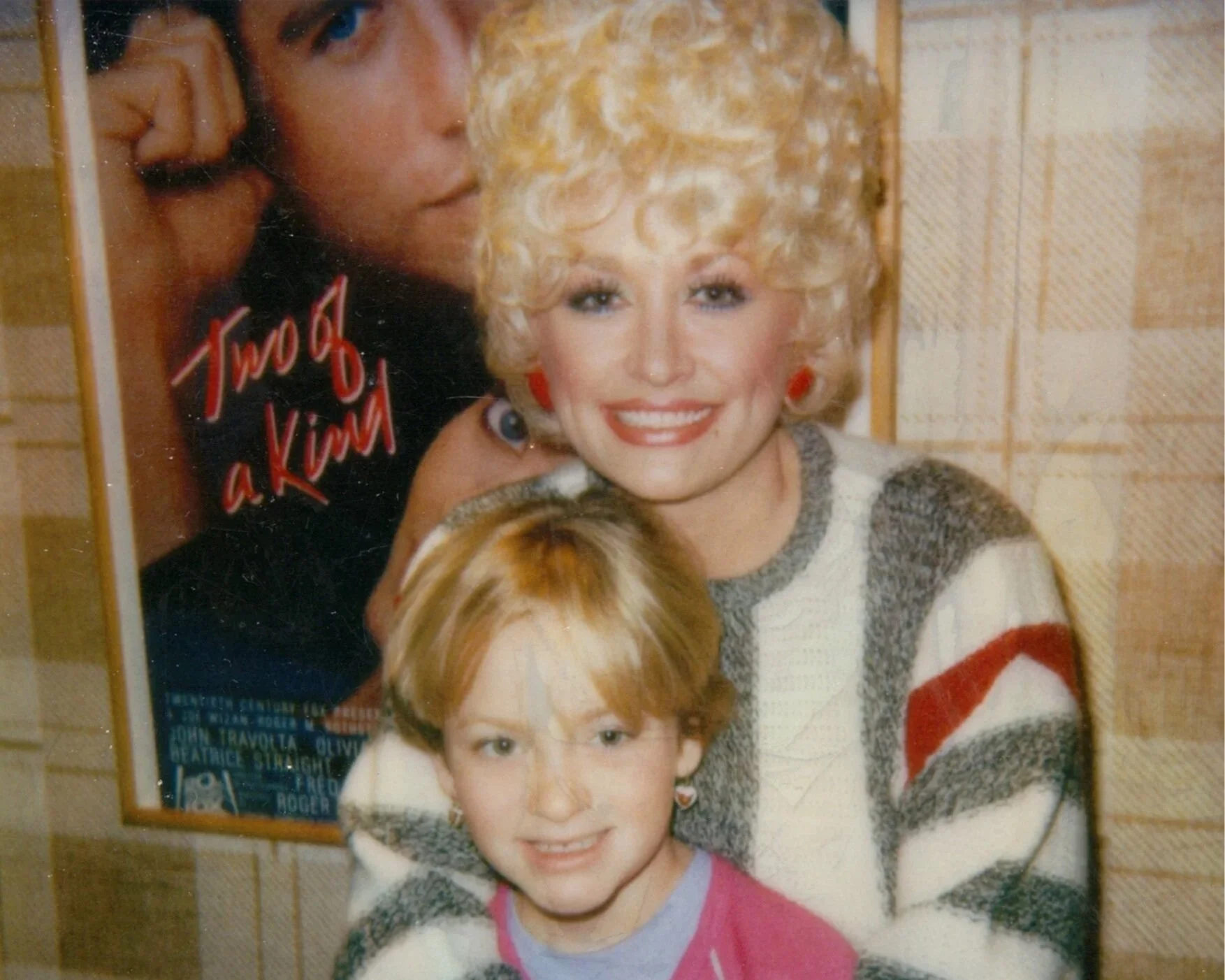 A vintage photograph featuring two women and two children. The woman in the background has blonde, curly hair, and is smiling, wearing a sweater with a striped pattern in red, white, gray, and black. The woman in front has straight, light brown hair, with a youthful face, wearing a jacket over a pink shirt. The children are positioned between the women; one has light blonde hair, and the other has dark hair, both with light skin. The background features a tiled wall with a poster or magazine page with red and black text, partially visible.
