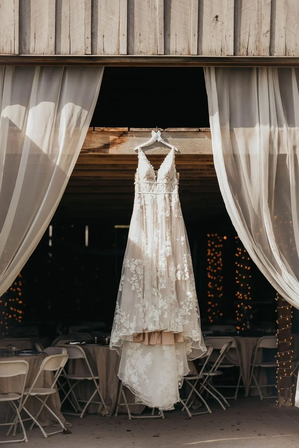 Lace wedding gown hanging in the barn doorway with string lights behind