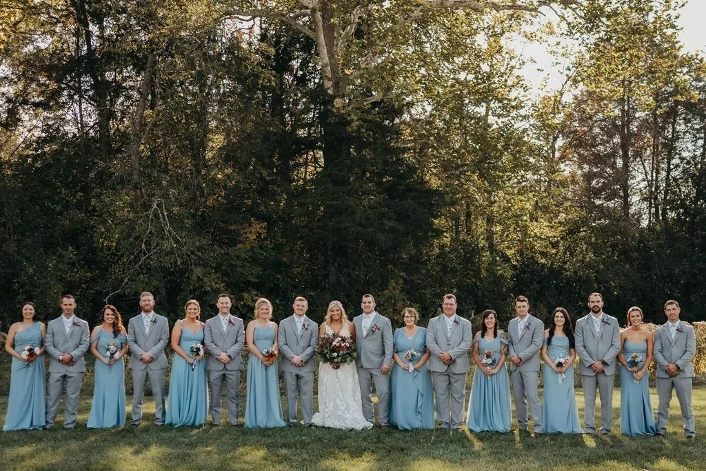 Wedding party in light blue and gray lined up in front of the tree line
