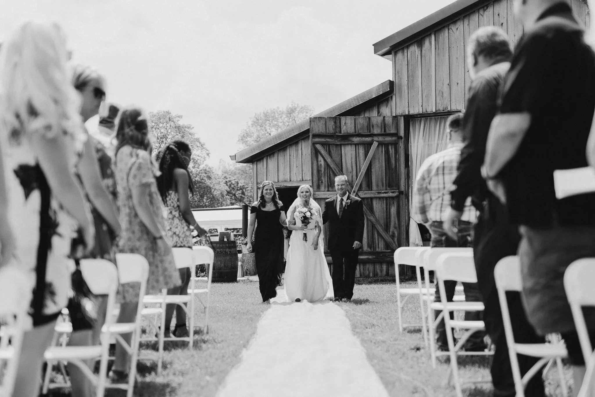 Bride walking down a white aisle runner toward the barn in black and white