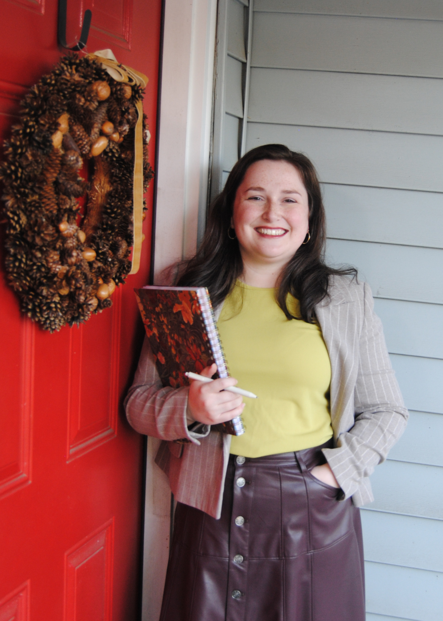 Smiling woman holding a floral notebook and pen beside a red door with a pinecone wreath