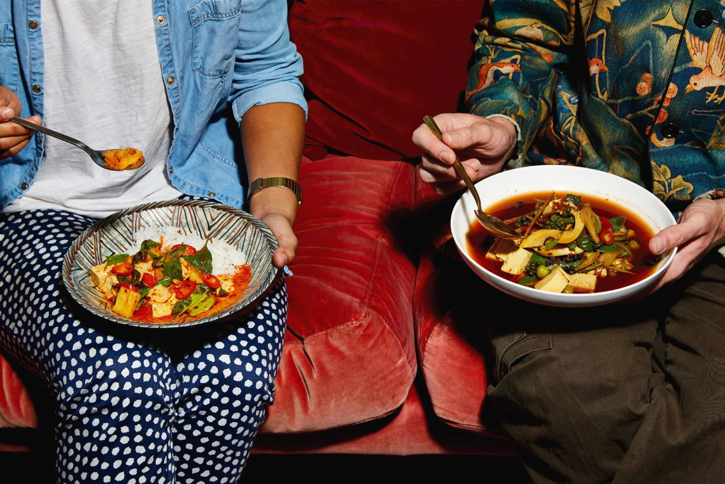 Two people eating colorful vegetable dishes with spoons while seated on a red couch.