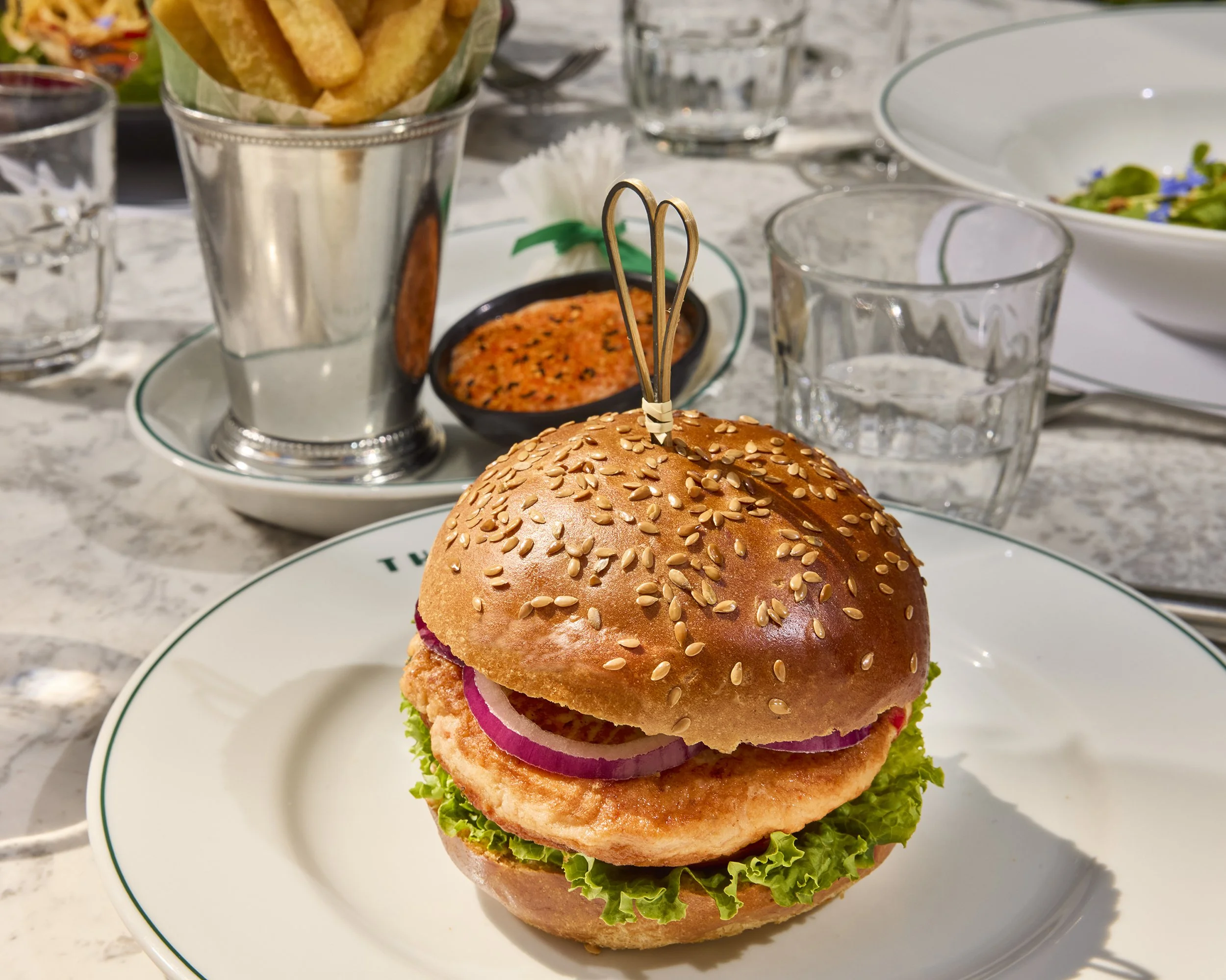 Gourmet chicken burger with lettuce and red onion on a sesame-seed bun, served with fries and dipping sauce.