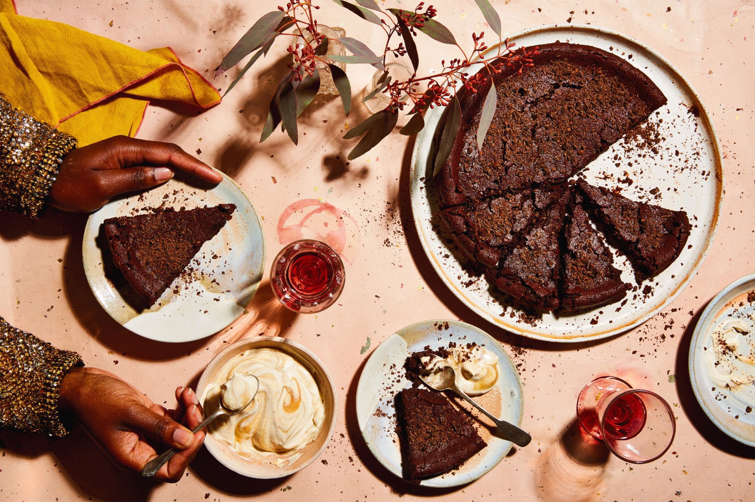 Overhead view of chocolate cake slices, whipped cream, red drinks, and hands serving dessert on a pink table