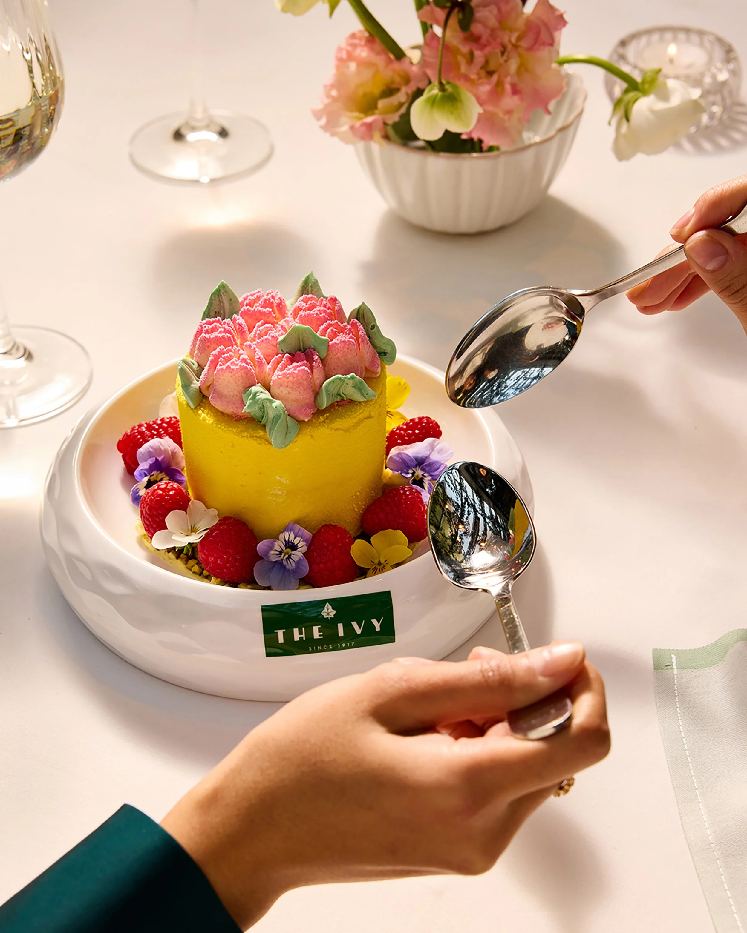 Yellow cake topped with pink frosting flowers, raspberries, and edible flowers on a white plate, with spoons and a floral centerpiece.