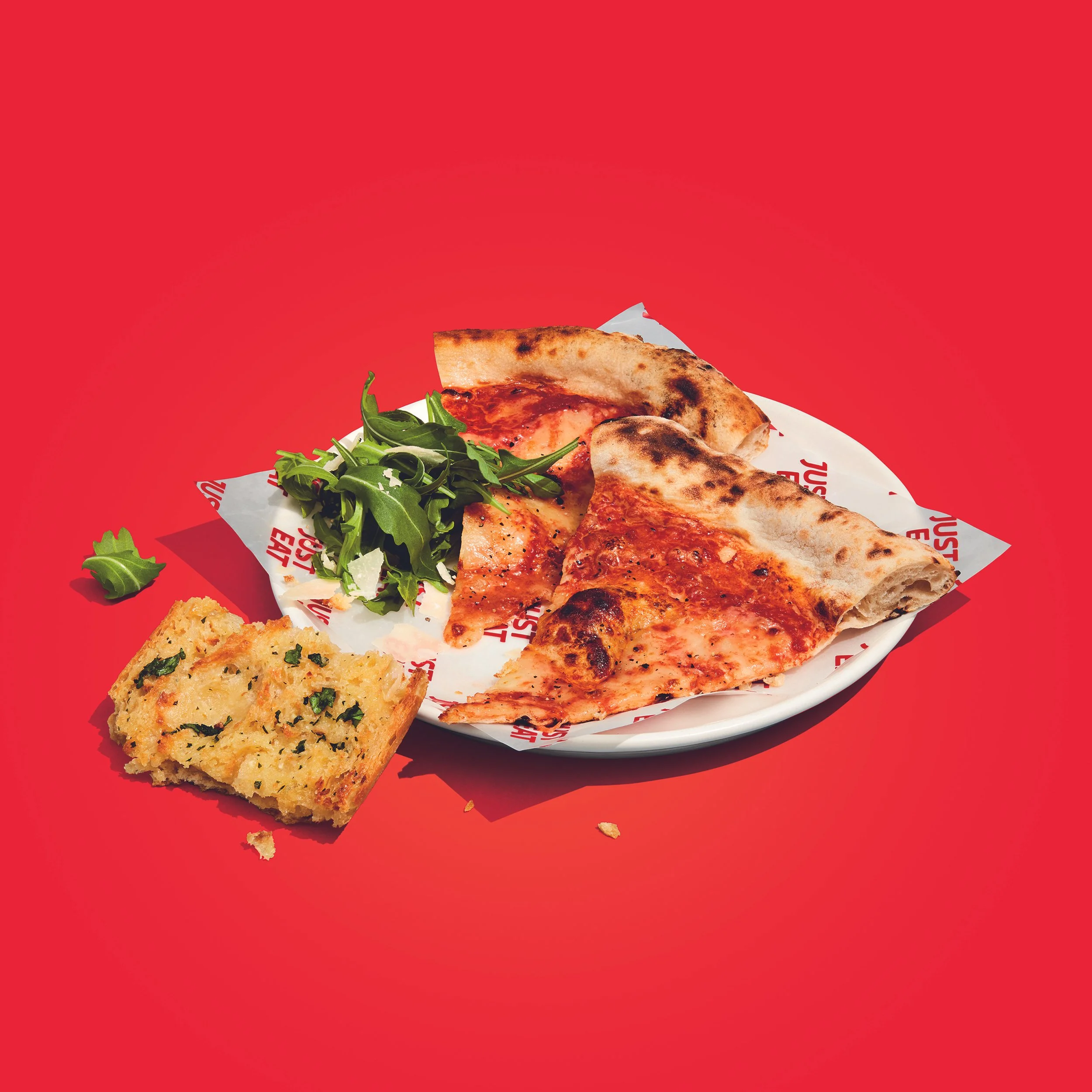 Sliced wood-fired Margherita pizza with arugula salad and garlic bread on a plate against a red background