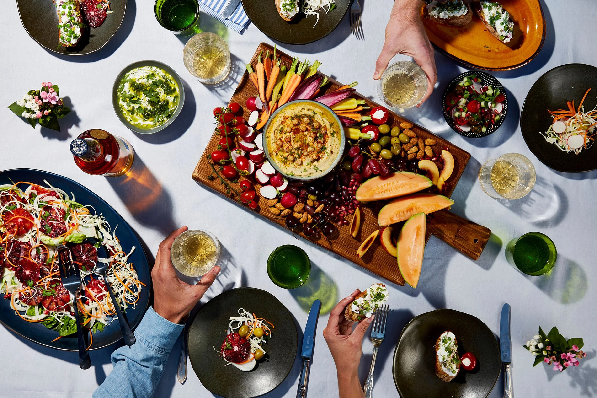 Overhead view of a festive table with hummus, colorful vegetables, fruit, salads, appetizers, wine, and diners sharing a meal