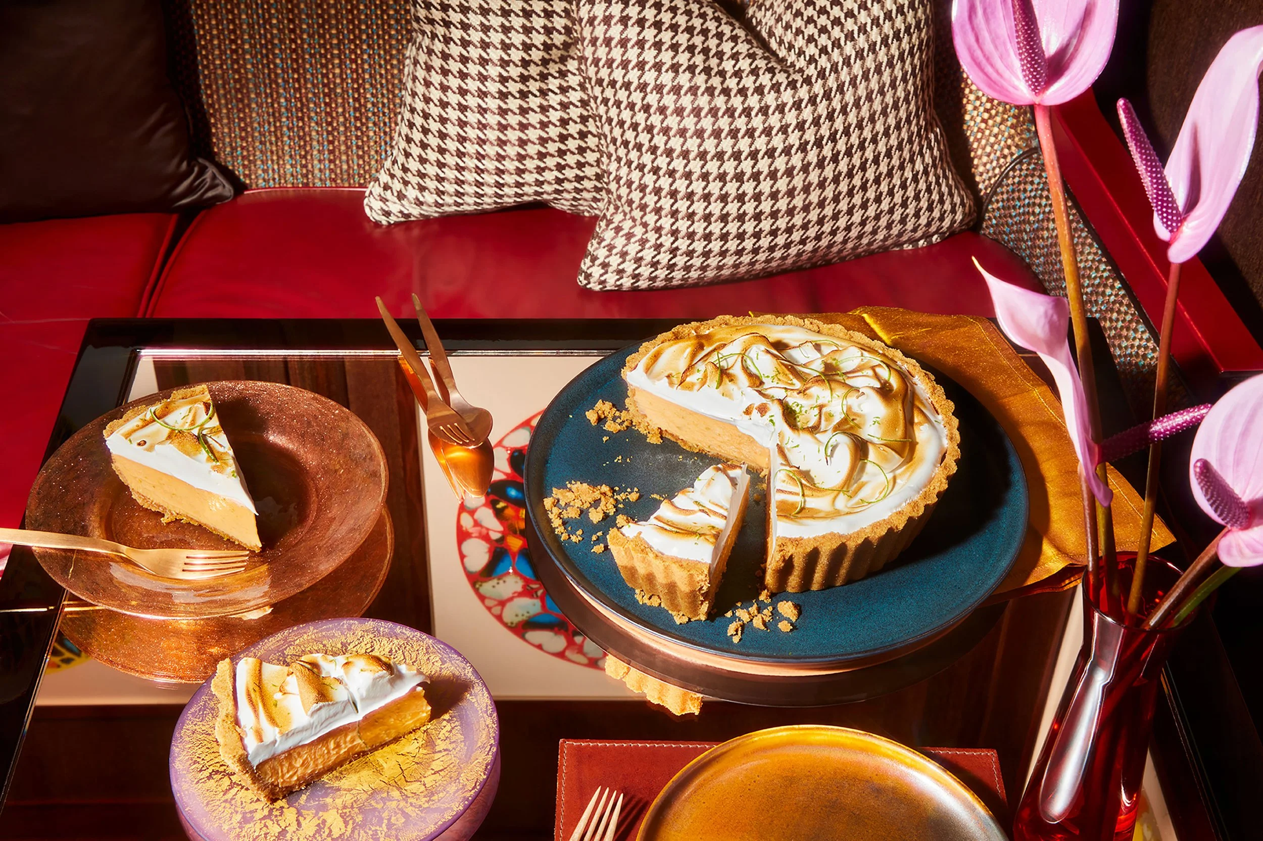 Slices of creamy citrus tart with toasted meringue on colorful plates beside a whole tart, gold flatware, pink flowers, and patterned cushions
