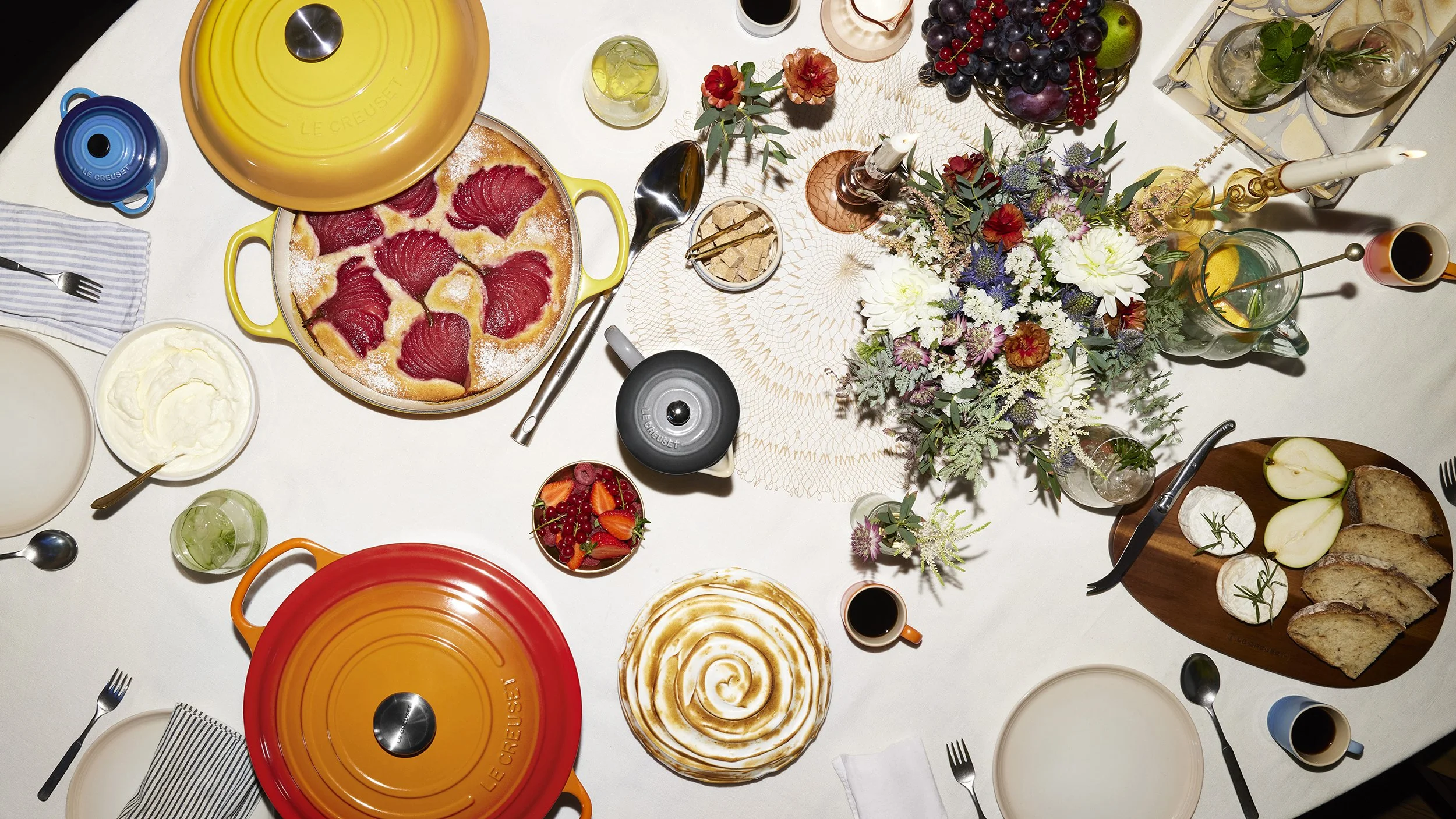 Overhead holiday table spread with pear tart, fruit, flowers, cheese, bread, cocktails, coffee, and colorful cookware