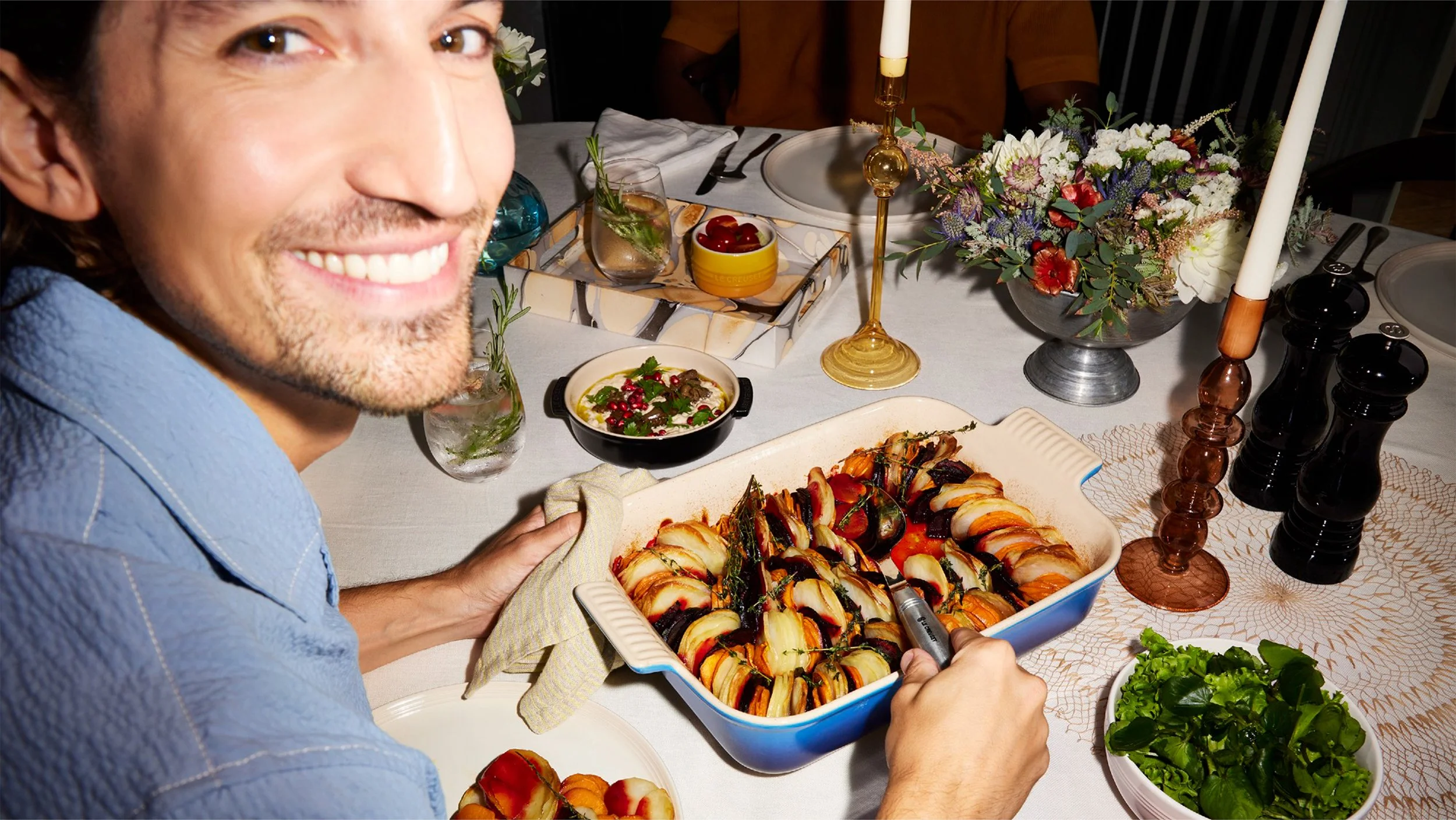 Smiling man serving a colorful roasted vegetable dish at a candlelit dinner table with flowers, salad, and appetizers