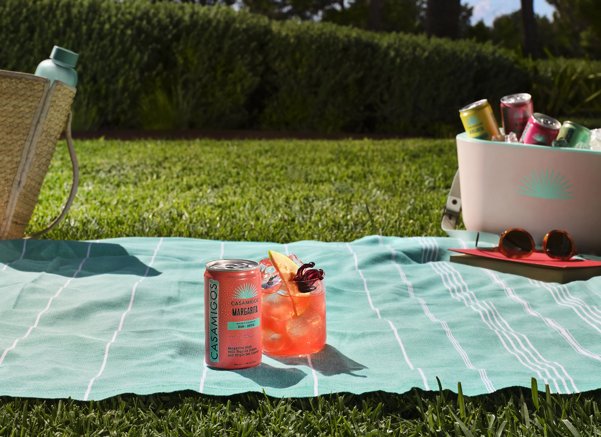 Outdoor picnic setup with a Casamigos Margarita can, garnished cocktail, colorful drinks in a cooler, sunglasses, and picnic blanket on a sunny lawn