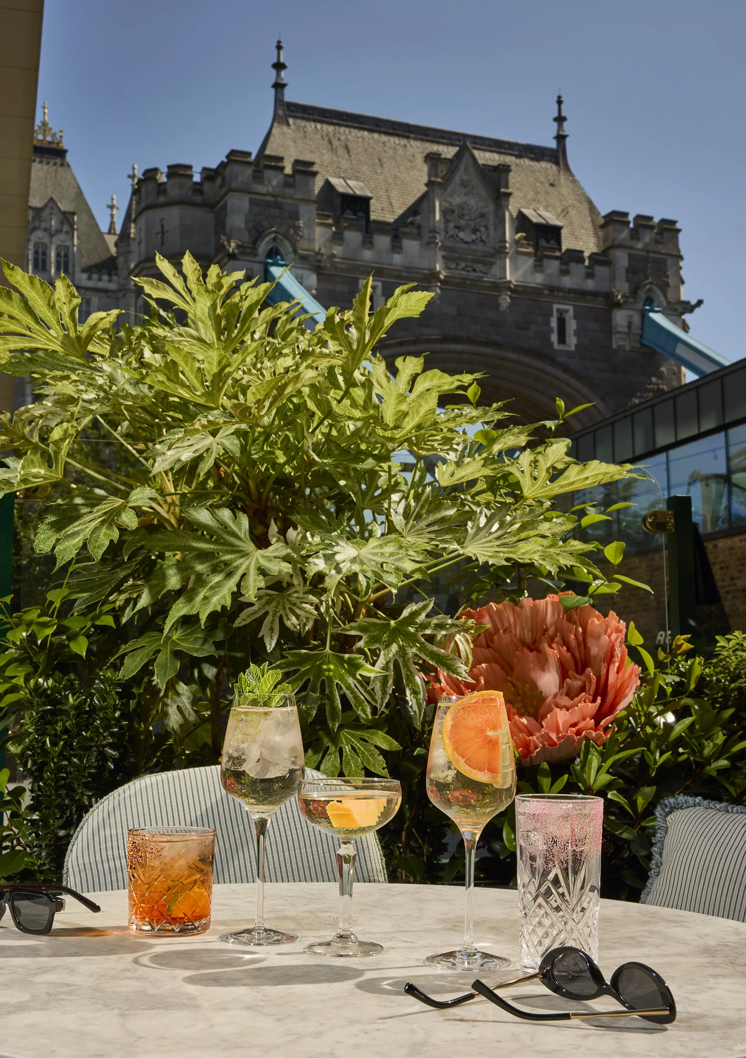 Outdoor dining table with colorful cocktails, citrus garnishes, sunglasses, lush greenery, and a historic Gothic building in the background