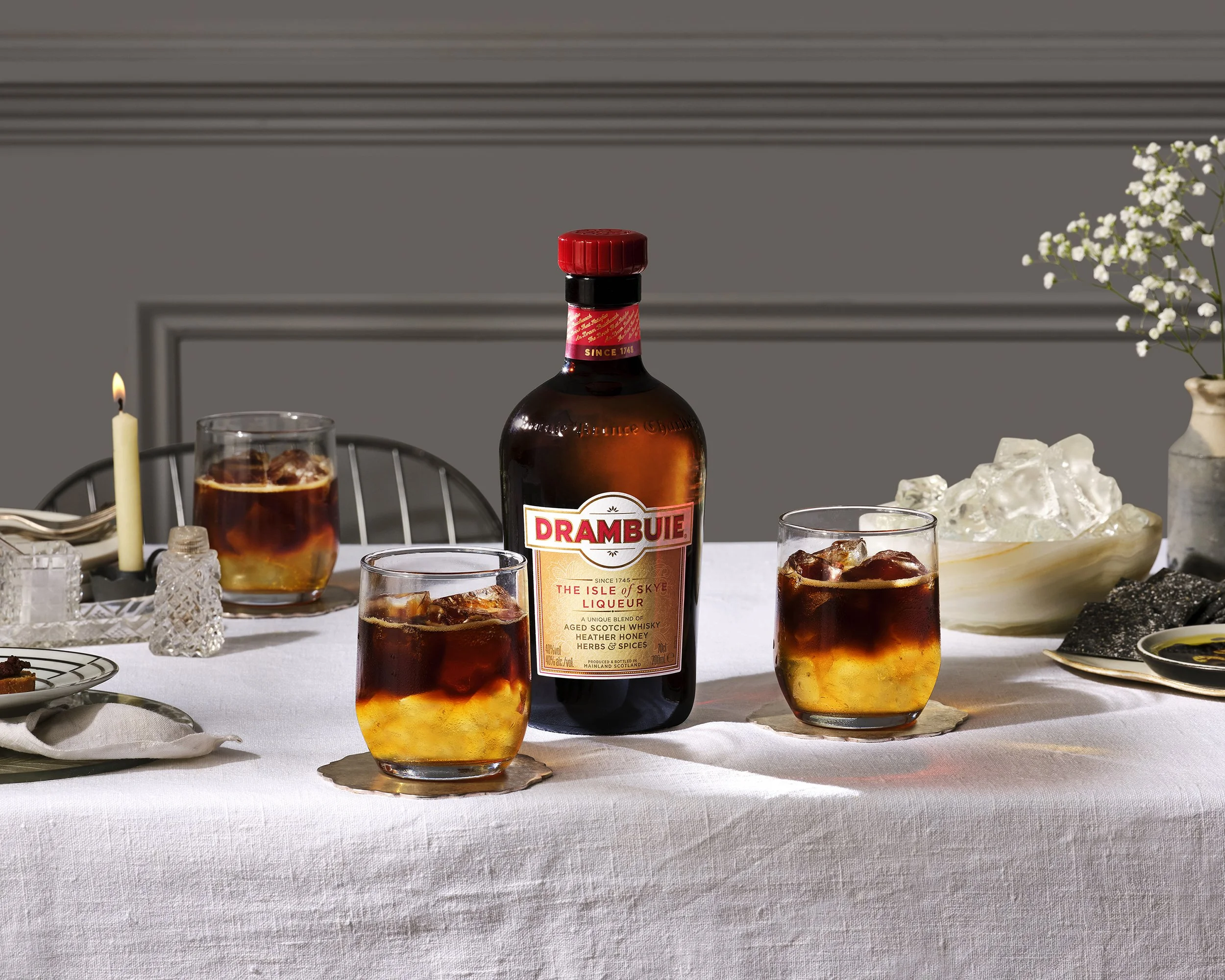 Drambuie Scotch whisky liqueur bottle surrounded by three whiskey cocktails with ice on a styled dining table
