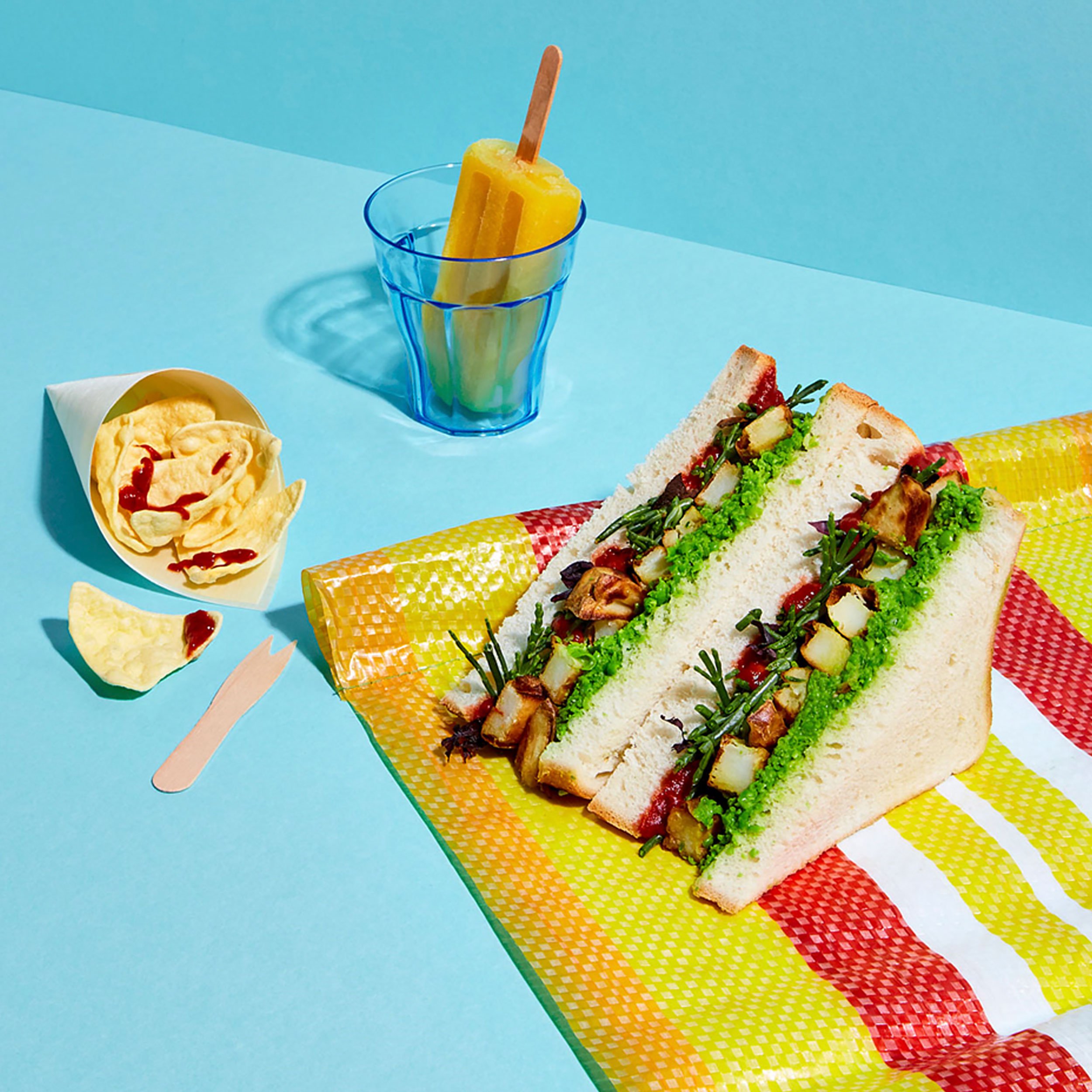 Colorful picnic spread with a vegetable sandwich, hummus and pita chips, and a yellow popsicle in a glass on a patterned blanket