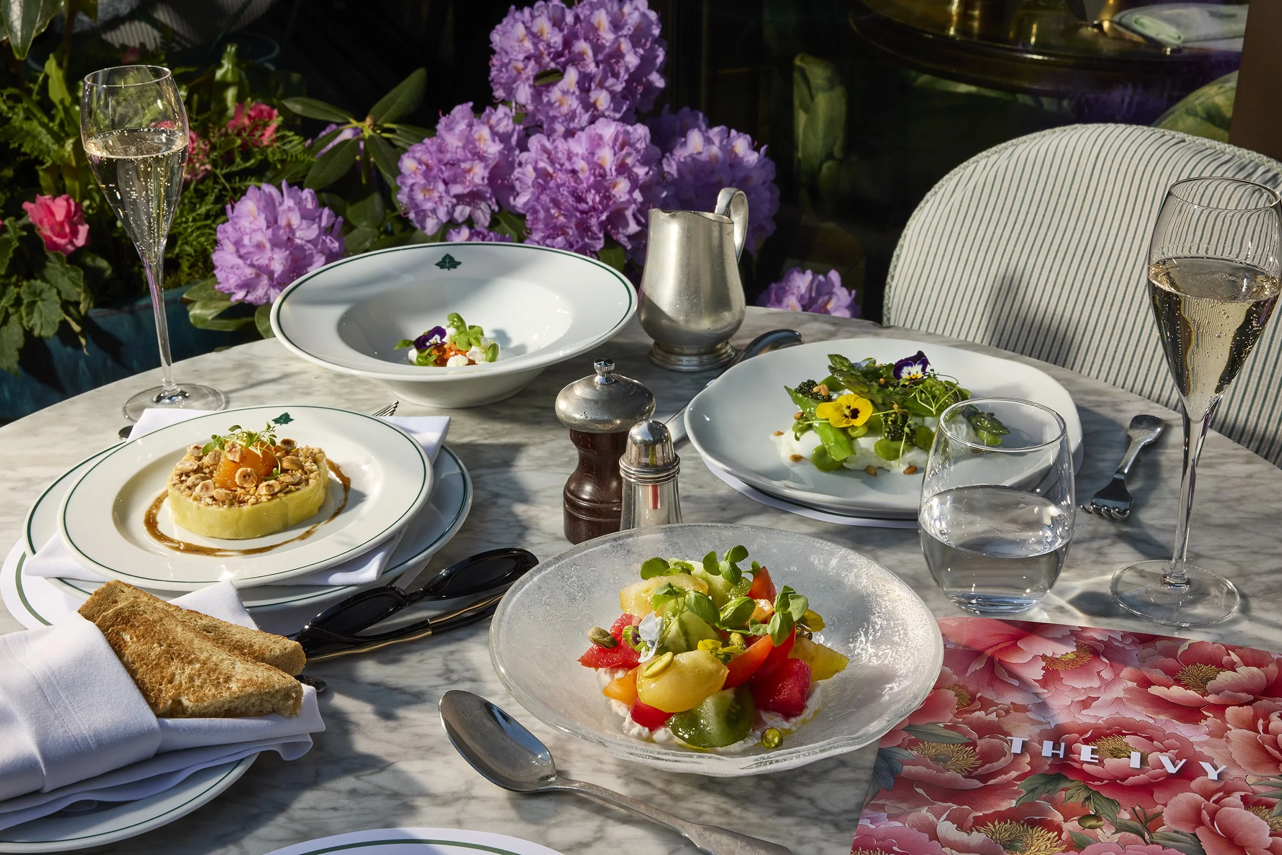 Elegant outdoor restaurant table with plated gourmet dishes, floral salads, champagne, wine glasses, and purple flowers