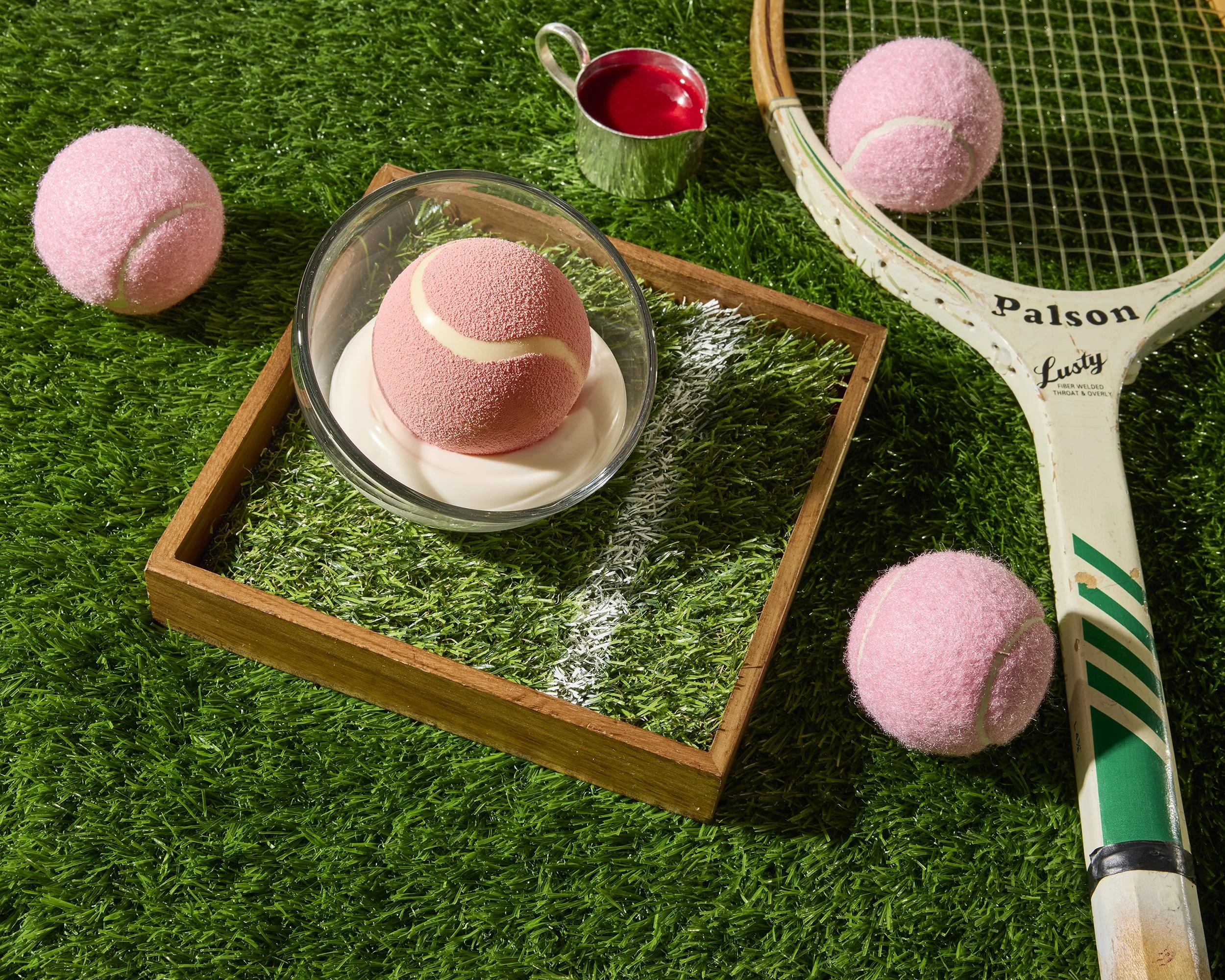 Pink tennis ball ice cream in a glass bowl on artificial grass, surrounded by tennis balls, a racket, and a cup of red juice.