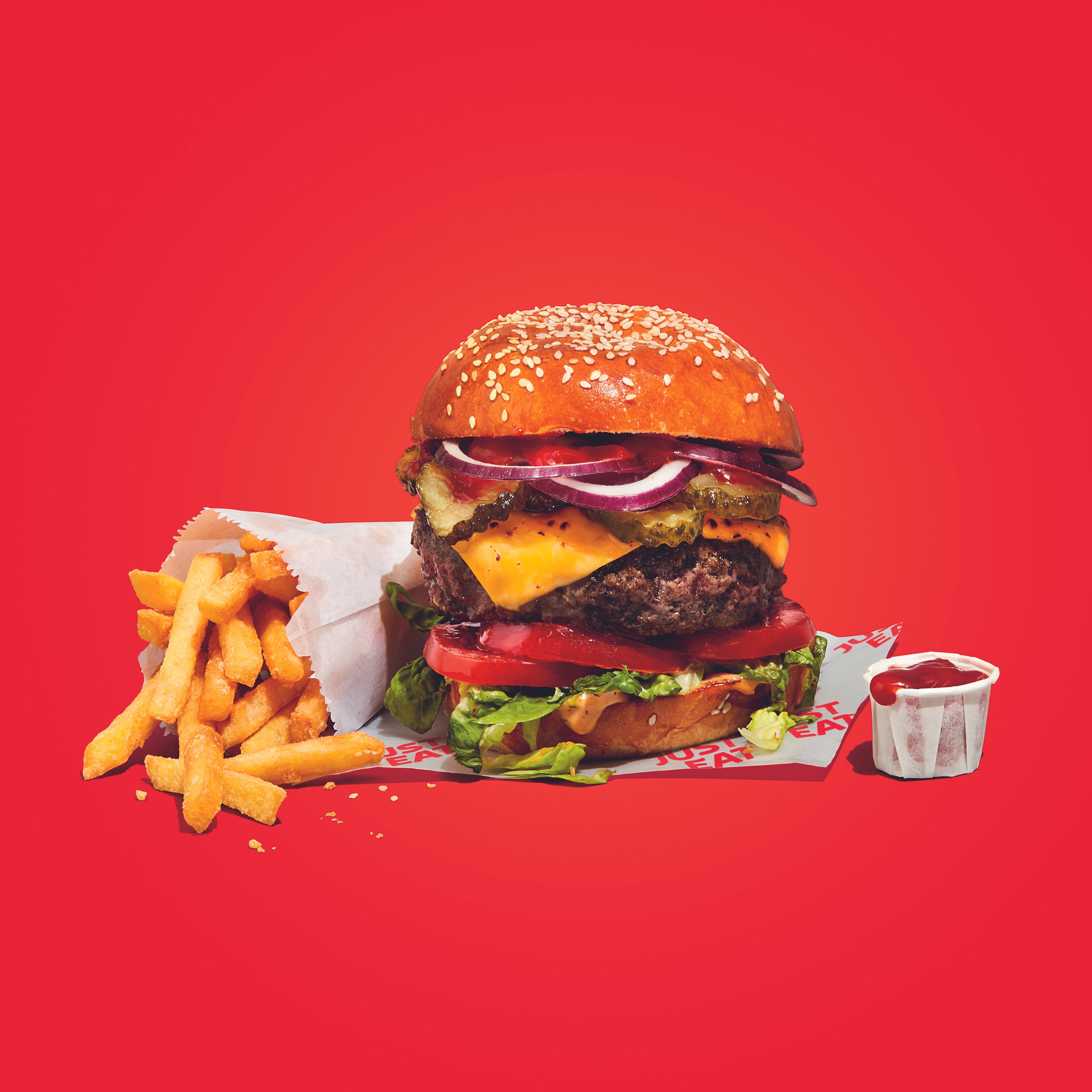 Double cheeseburger with sesame bun, lettuce, tomato, onions, pickles, and fries with ketchup on a red background