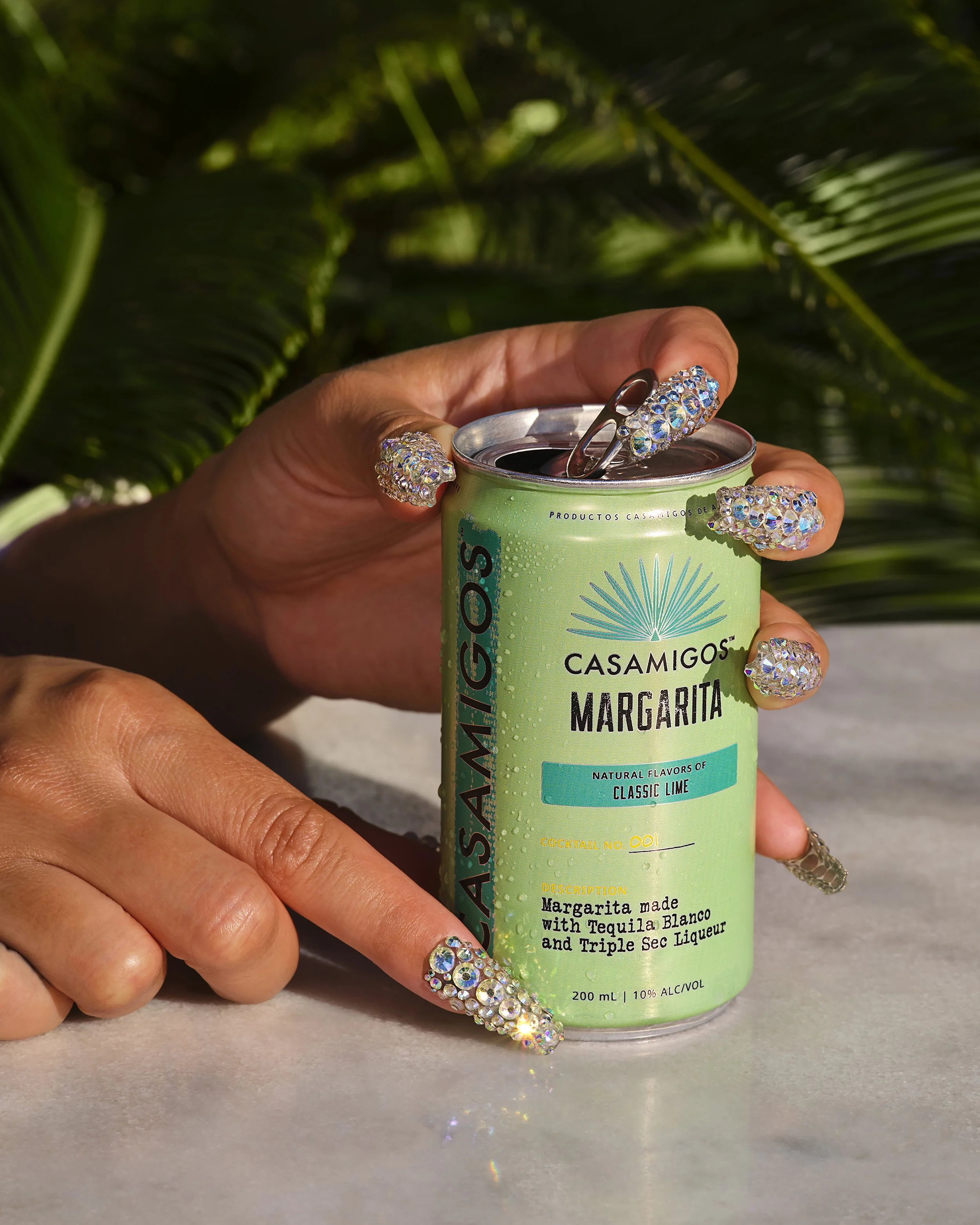 Hand with rhinestone-encrusted nails holding a Casamigos Classic Lime Margarita can against a leafy green background
