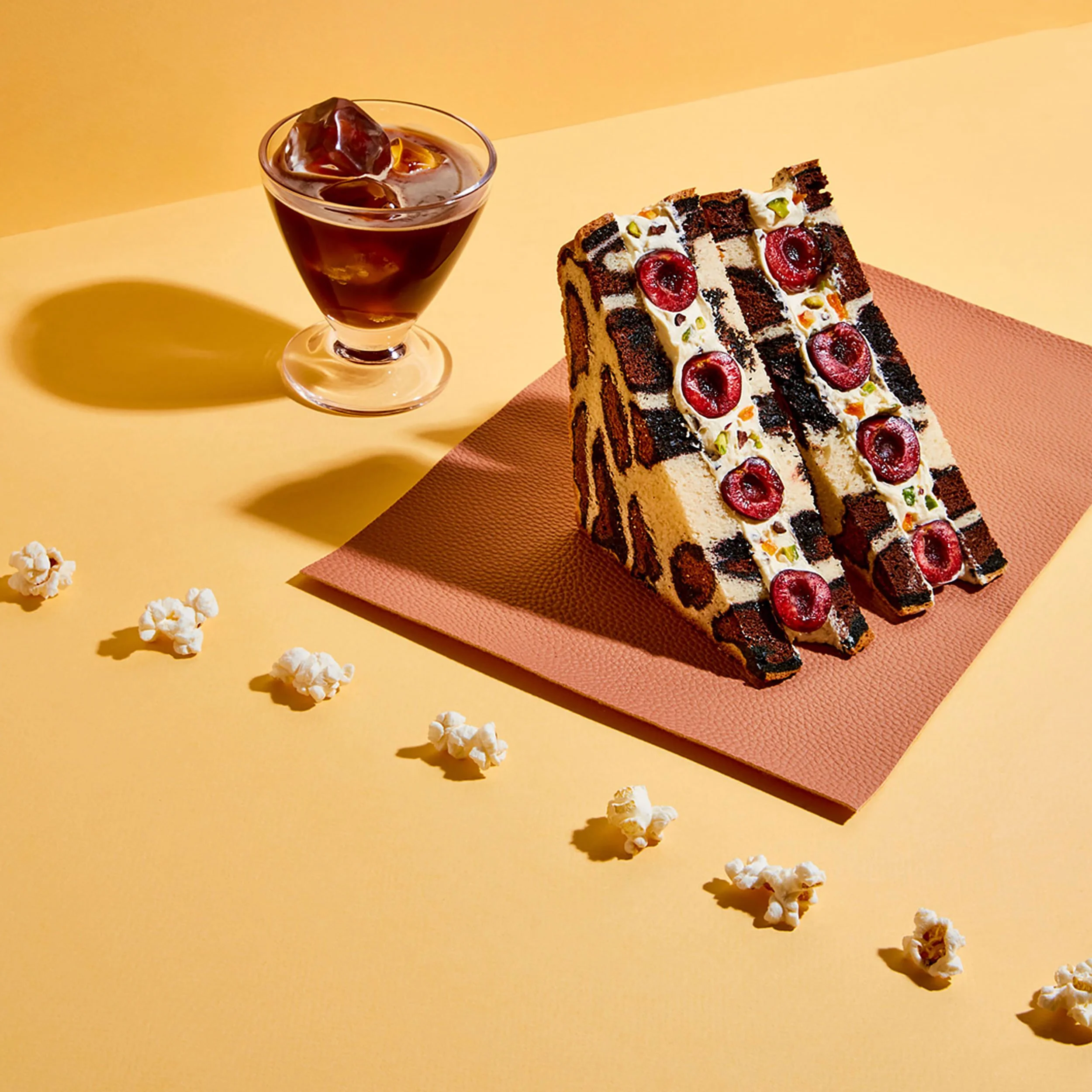 Chocolate cake topped with whipped cream, cherries, pistachios, and chocolate pieces beside an iced coffee and scattered popcorn.