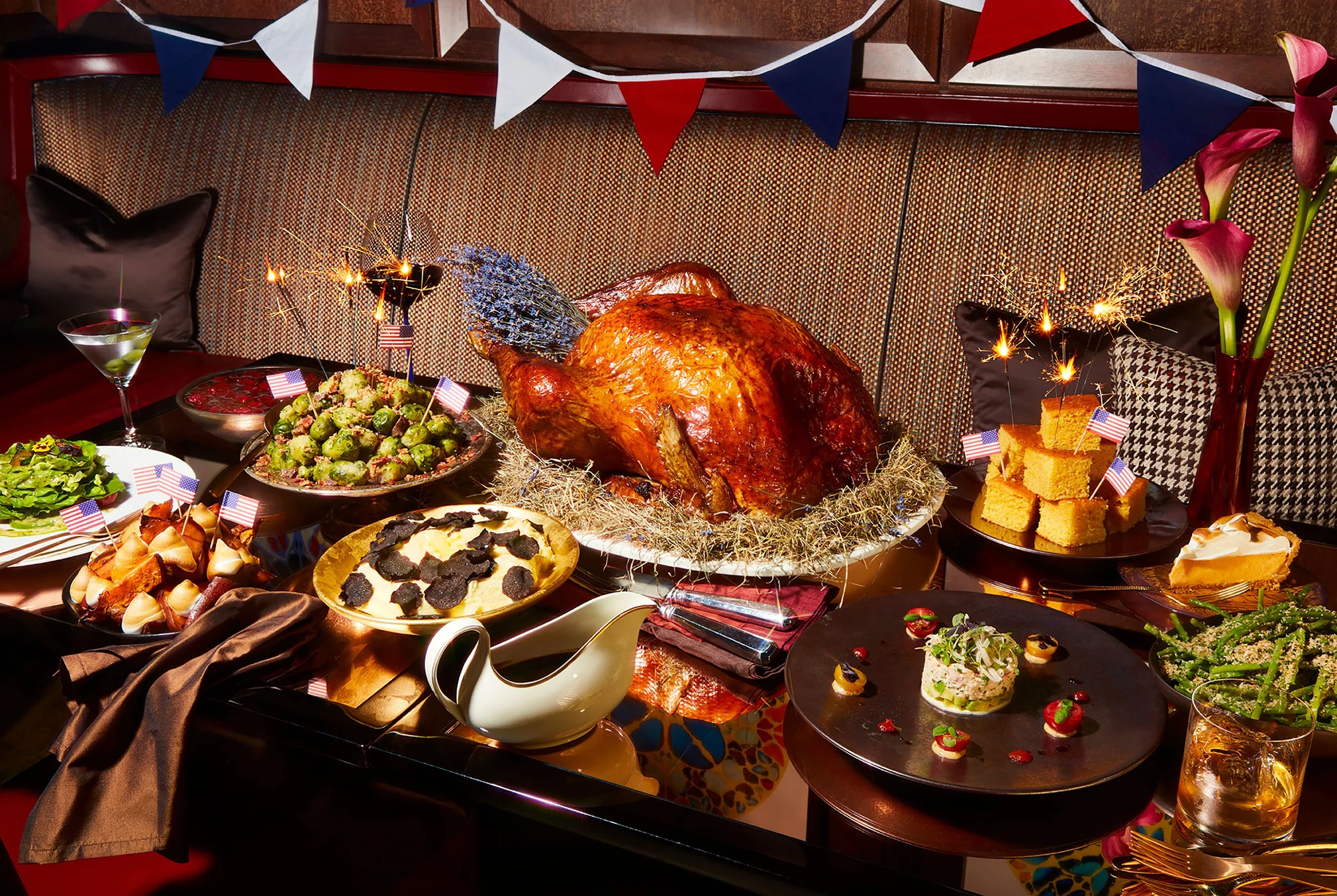 Festive Thanksgiving dinner spread with roasted turkey, side dishes, cornbread, pie, cocktails, sparklers, flowers, and American flag decorations
