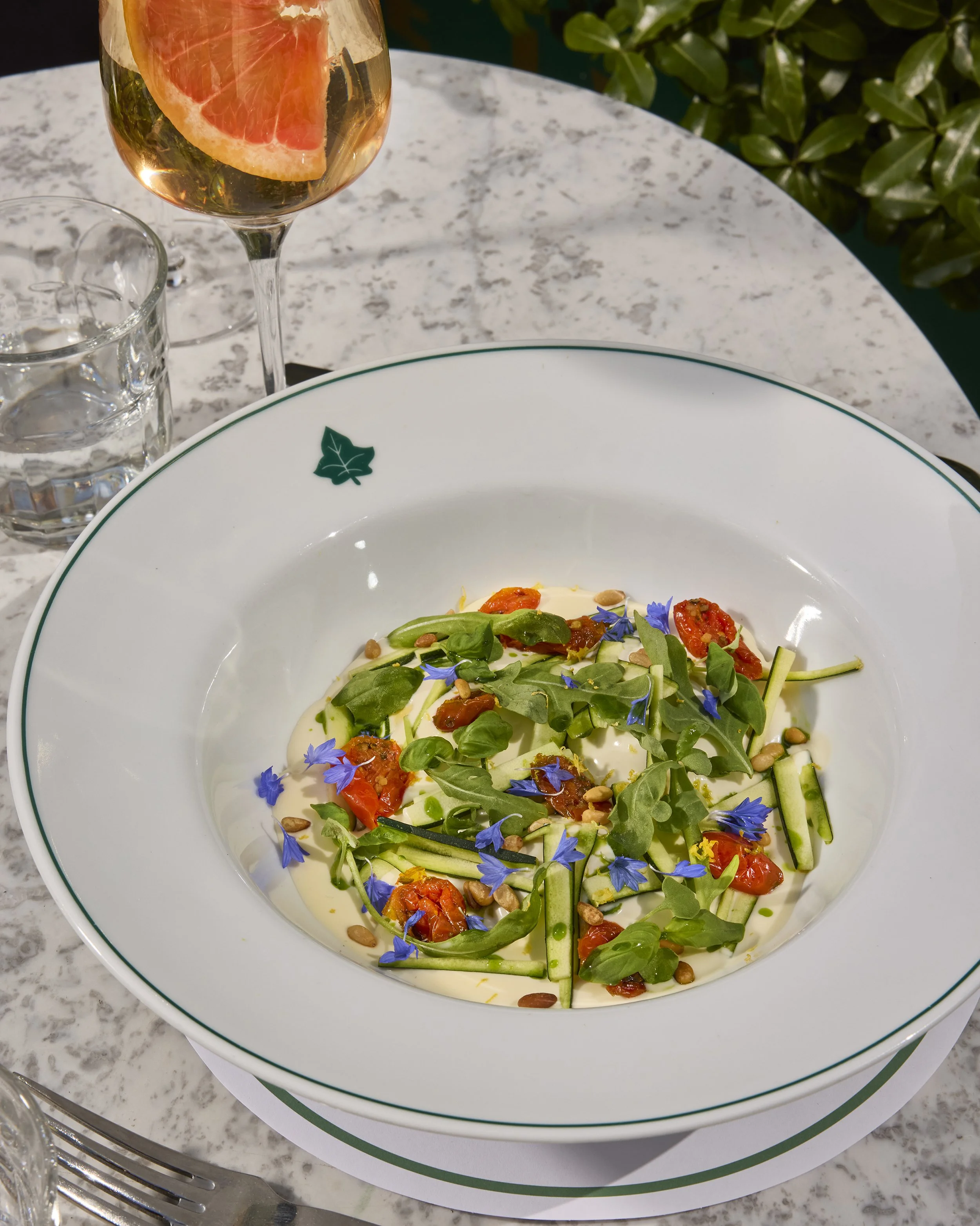 Gourmet salad with zucchini, roasted tomatoes, greens, pine nuts, and edible blue flowers beside a grapefruit cocktail