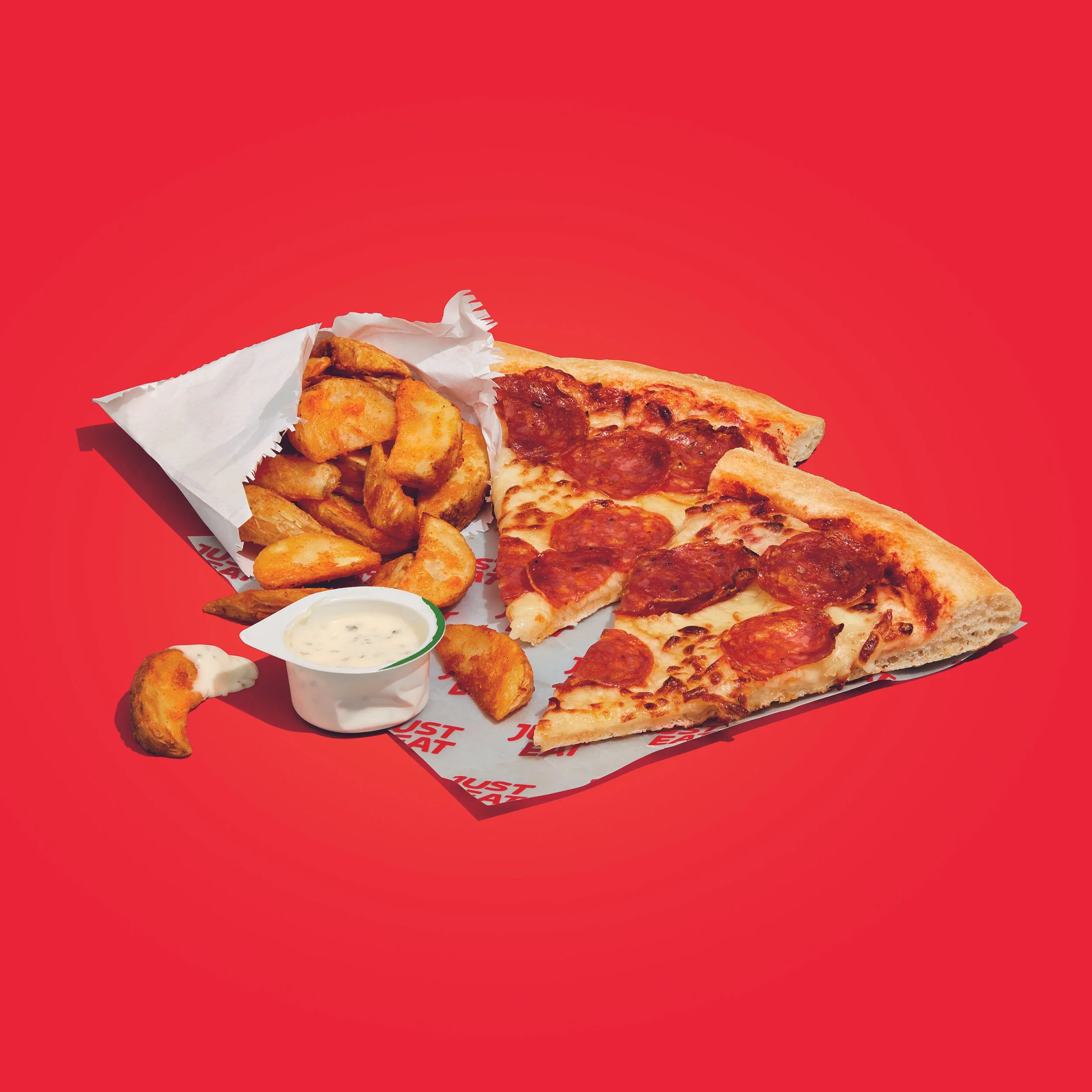 Pepperoni pizza slices with seasoned potato wedges and ranch dipping sauce on a red background