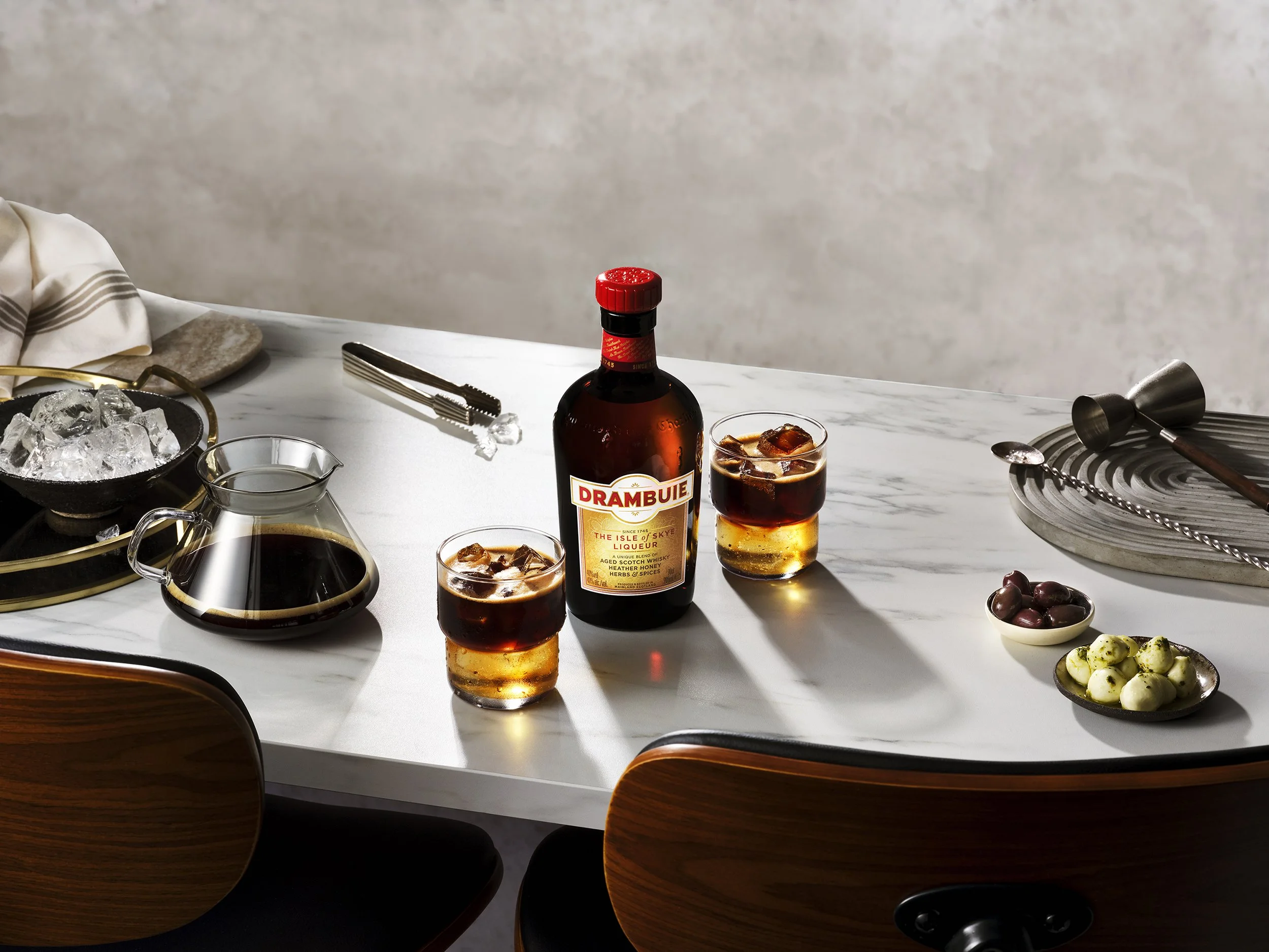 Drambuie liqueur bottle with two iced cocktails on a marble table, surrounded by coffee, ice, bar tools, olives, and snacks
