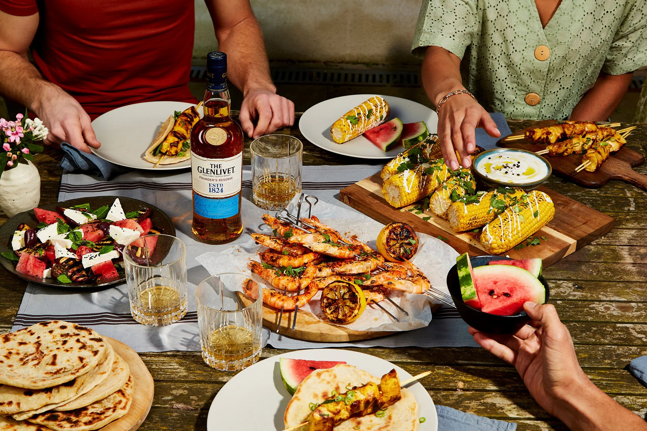 Outdoor barbecue feast with grilled shrimp, corn, skewers, flatbread, watermelon salad, and The Glenlivet whisky