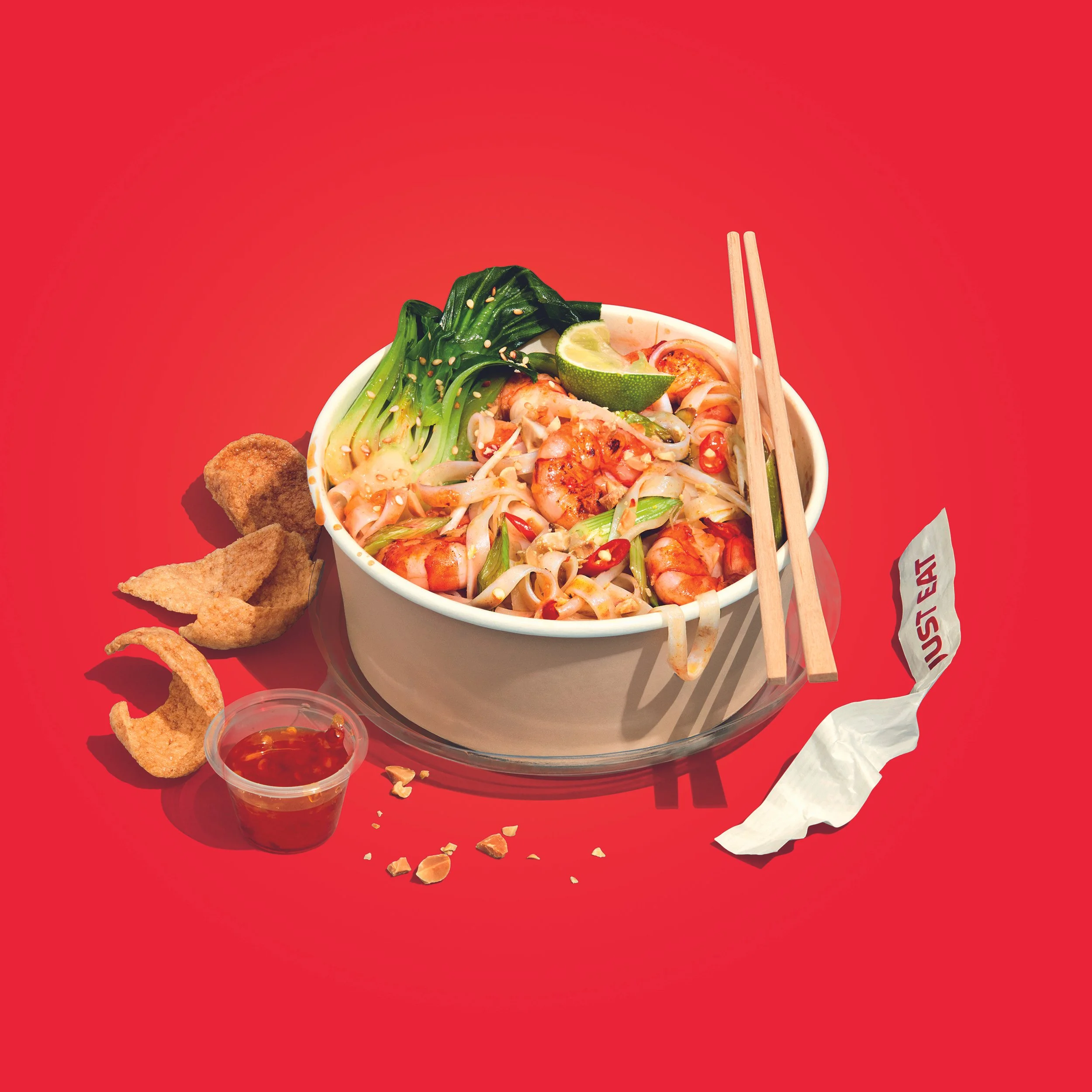 Bowl of shrimp noodles with vegetables, lime, chopsticks, fried appetizers, and dipping sauce on a red table